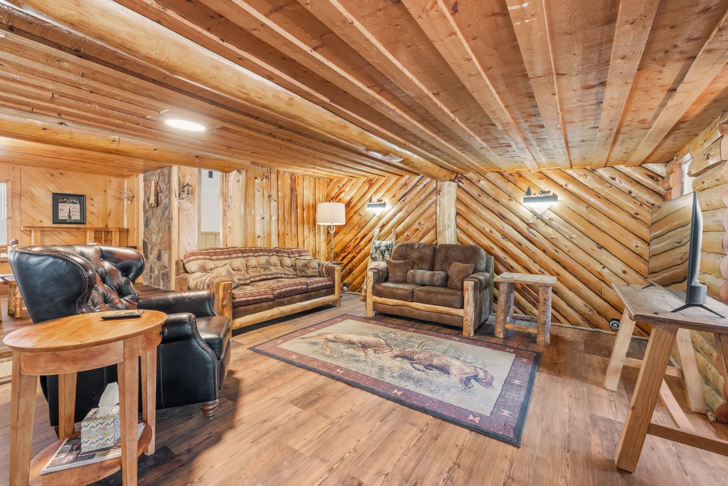 A rustic living room with log-paneled walls, exposed wood beam ceiling, wood-look flooring, sofas with log frames, and an armchair.