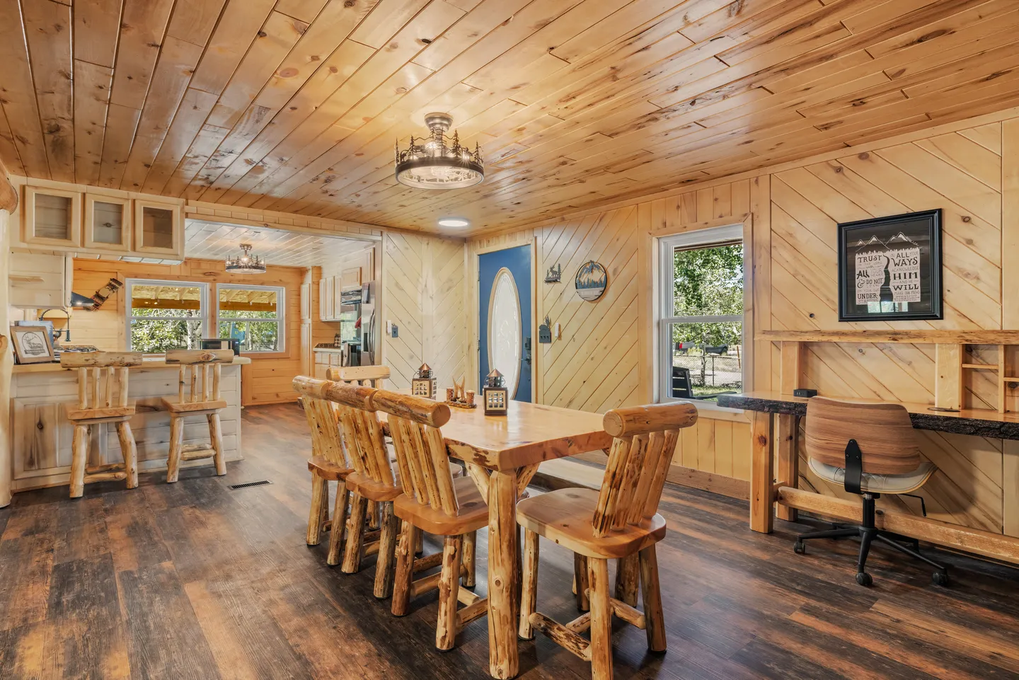 Dining area with rustic log table and chairs, wood-paneled walls and ceiling, looking toward an open kitchen and front door.
