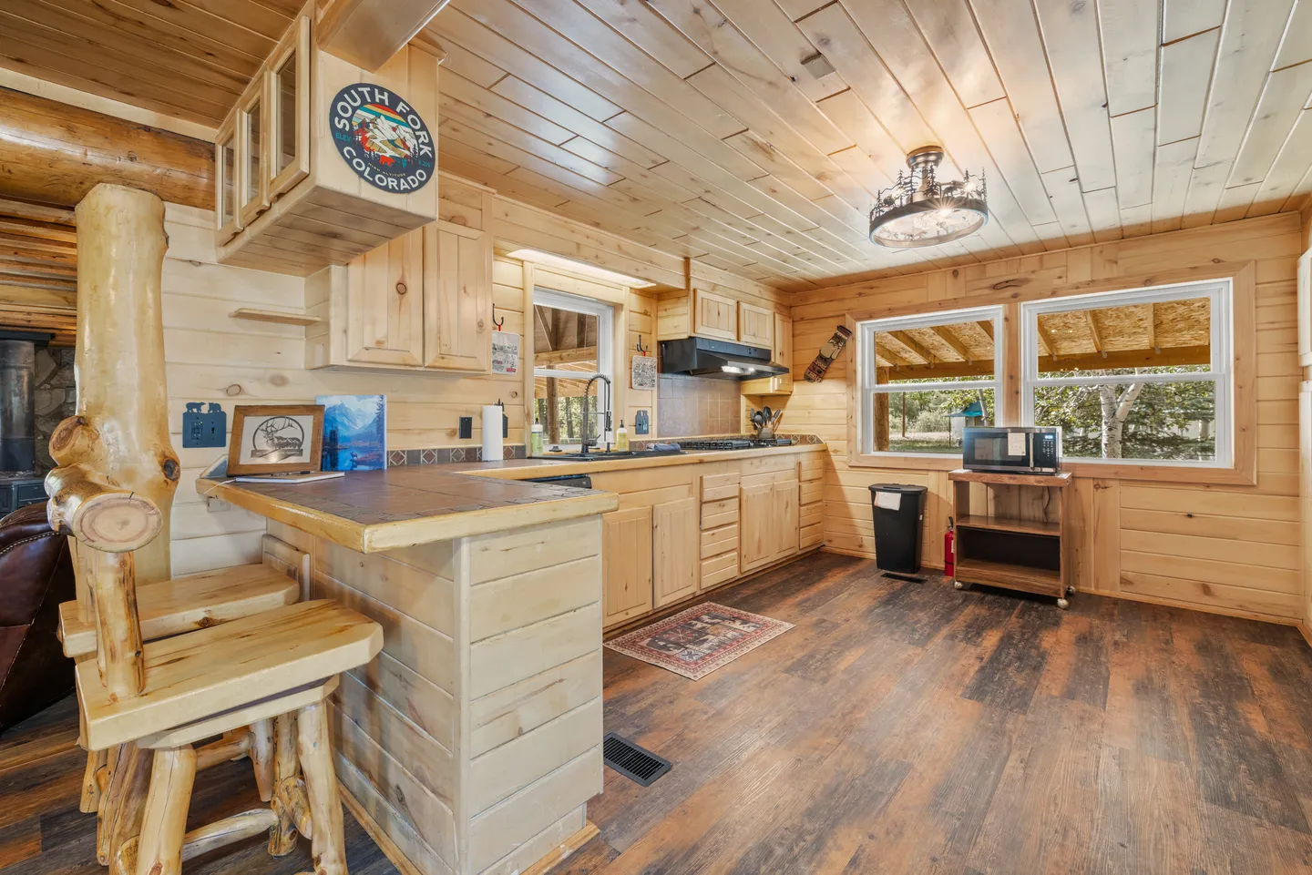 Kitchen in a log-style cabin with wood-paneled walls and ceiling, light wood cabinetry, and a small breakfast bar with log barstools.