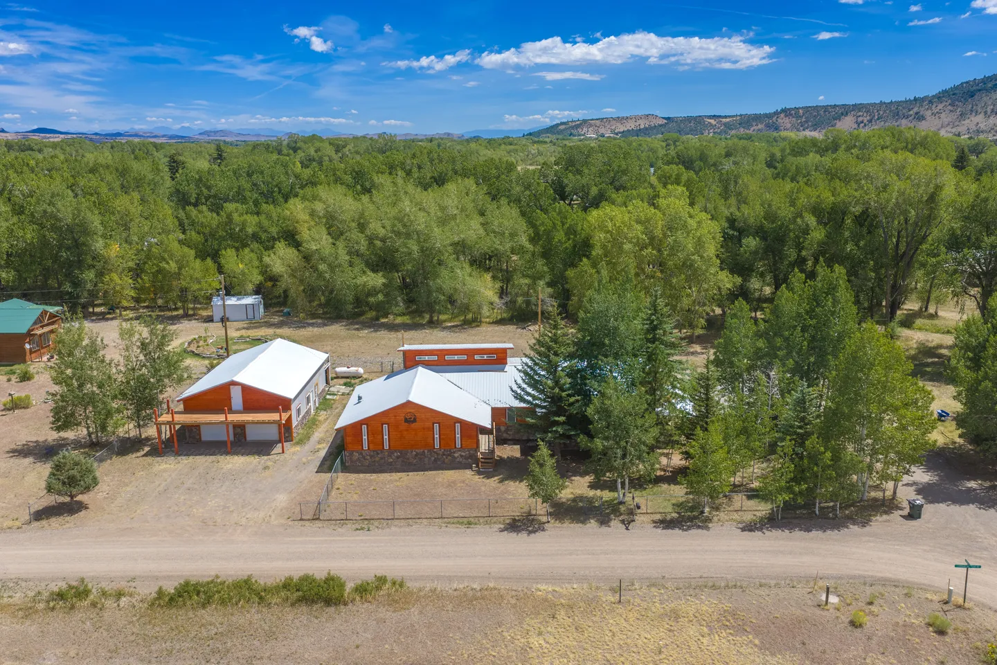 Aerial view of a rural property featuring multiple wood-sided buildings with metal roofs, surrounded by dense trees with hills in the background.