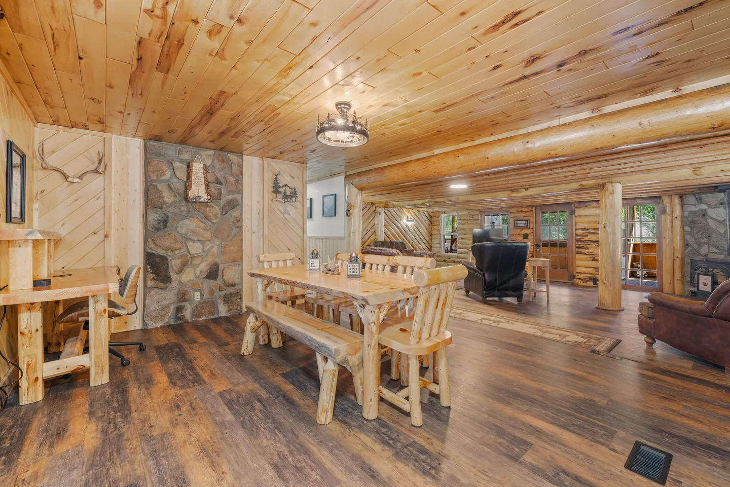 Open-concept cabin interior featuring a rustic log dining table with benches, wood plank ceilings, and stone wall accents.