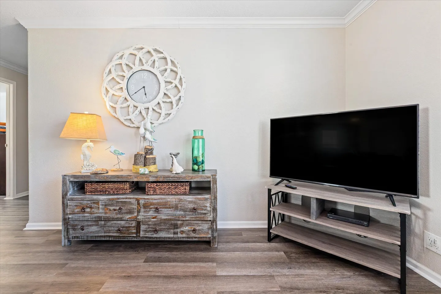 A living room corner with wood-look flooring, featuring a console table with coastal decor and a mounted ornate clock, beside a television on a media stand.