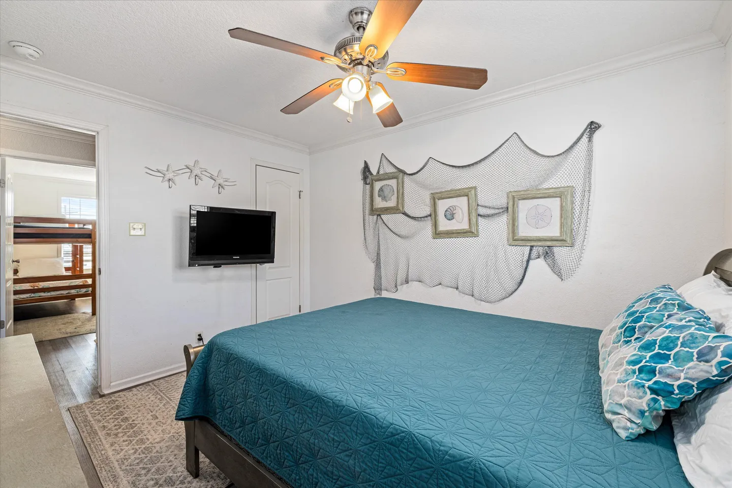 Bedroom featuring a bed with a teal coverlet, a wall-mounted TV, a ceiling fan, and coastal wall decor.