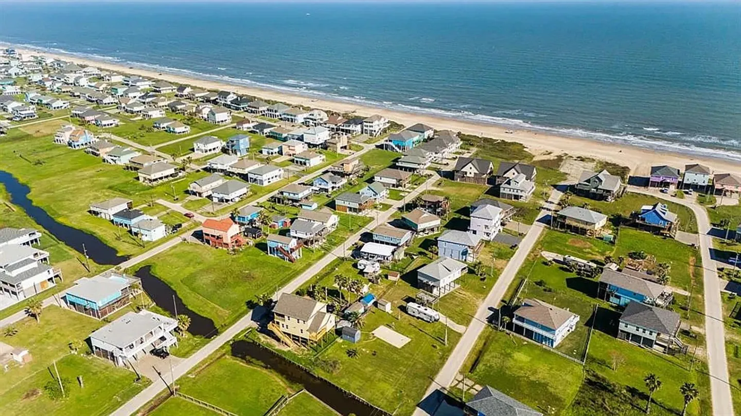 Aerial view of a coastal neighborhood with stilted beach houses along a sandy shoreline and ocean.