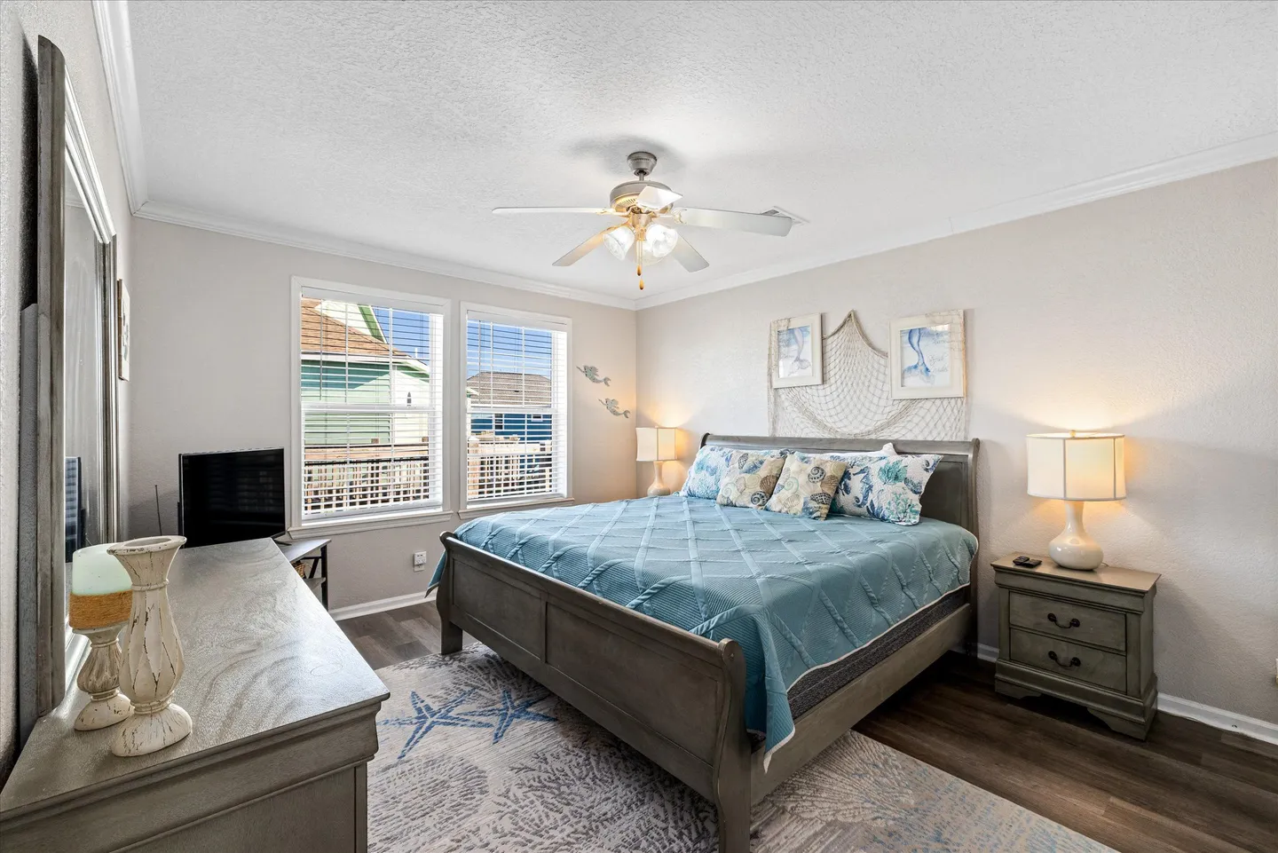 Bedroom with a gray wooden bed frame, blue quilt, two windows, nightstands with lamps, and a dresser.