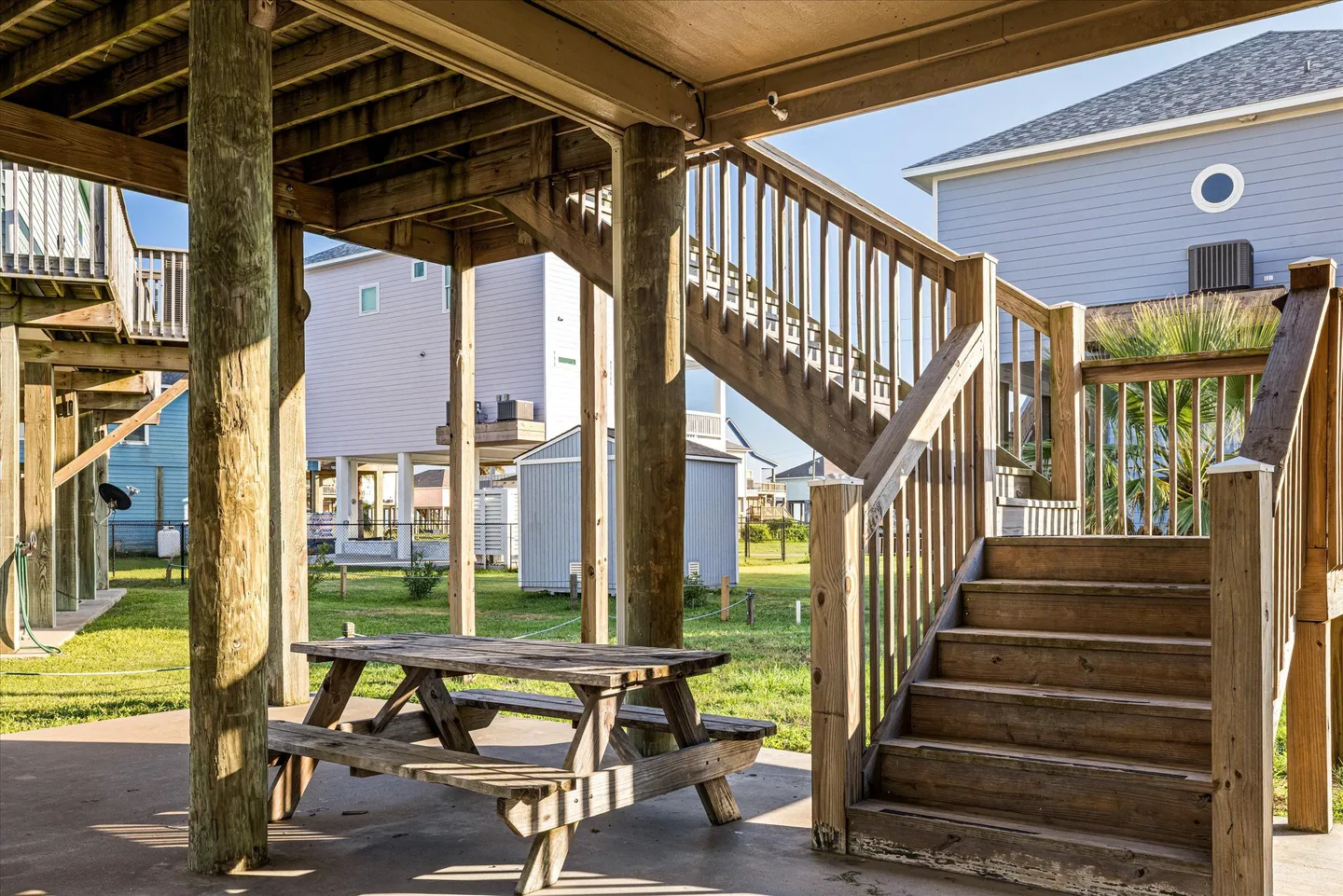 Covered ground-level patio beneath a stilt home, featuring a wooden picnic table and a wooden staircase leading up.
