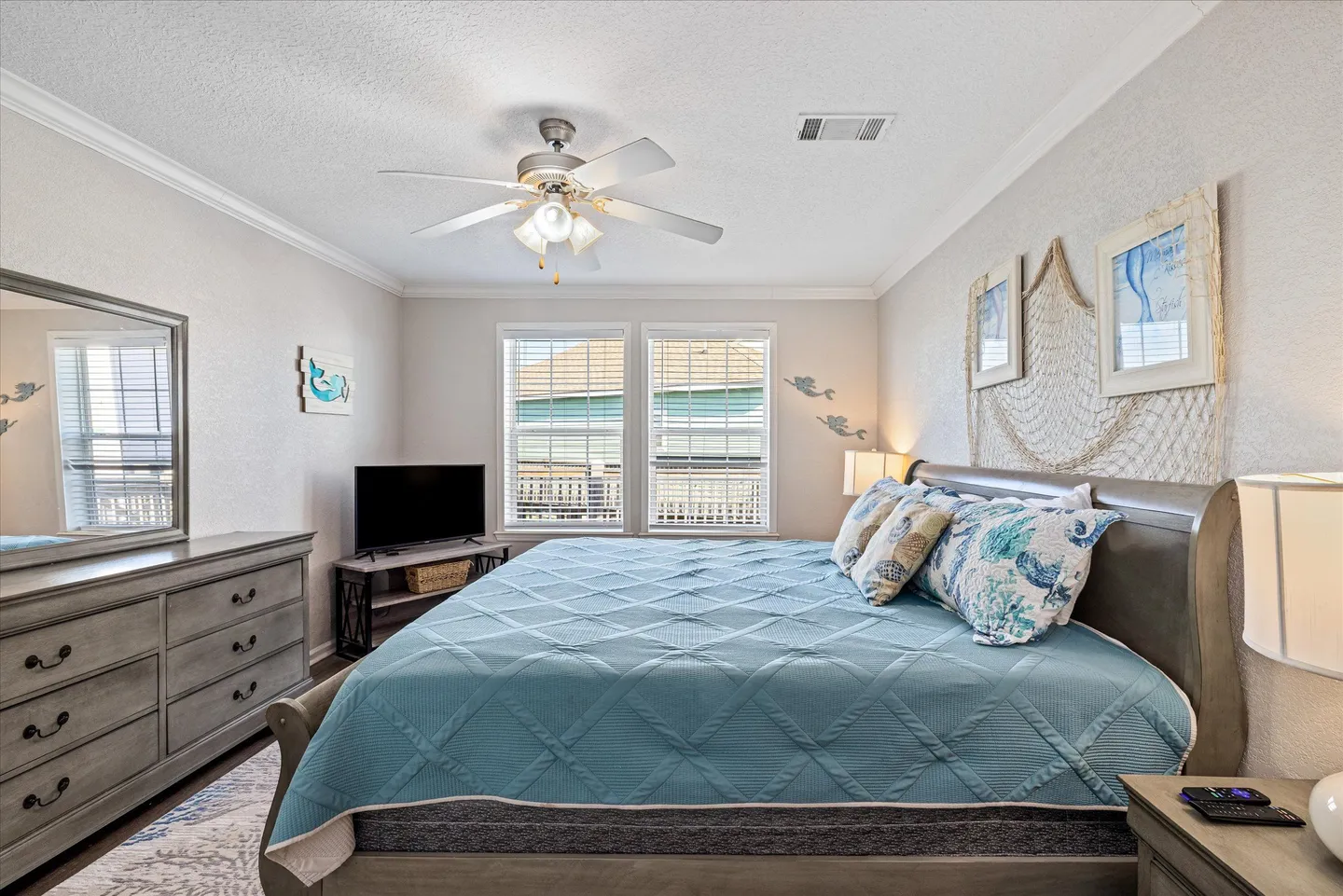 Bedroom with a large bed dressed in a blue quilt, matching wooden dresser with mirror, and coastal-themed wall decorations.