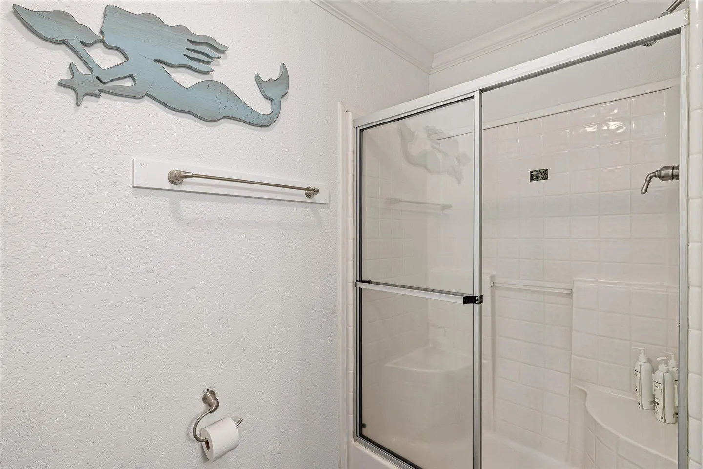 Bathroom showing a walk-in shower with sliding glass doors, built-in corner seats, a towel bar, toilet paper holder, and a wooden mermaid wall decoration.
