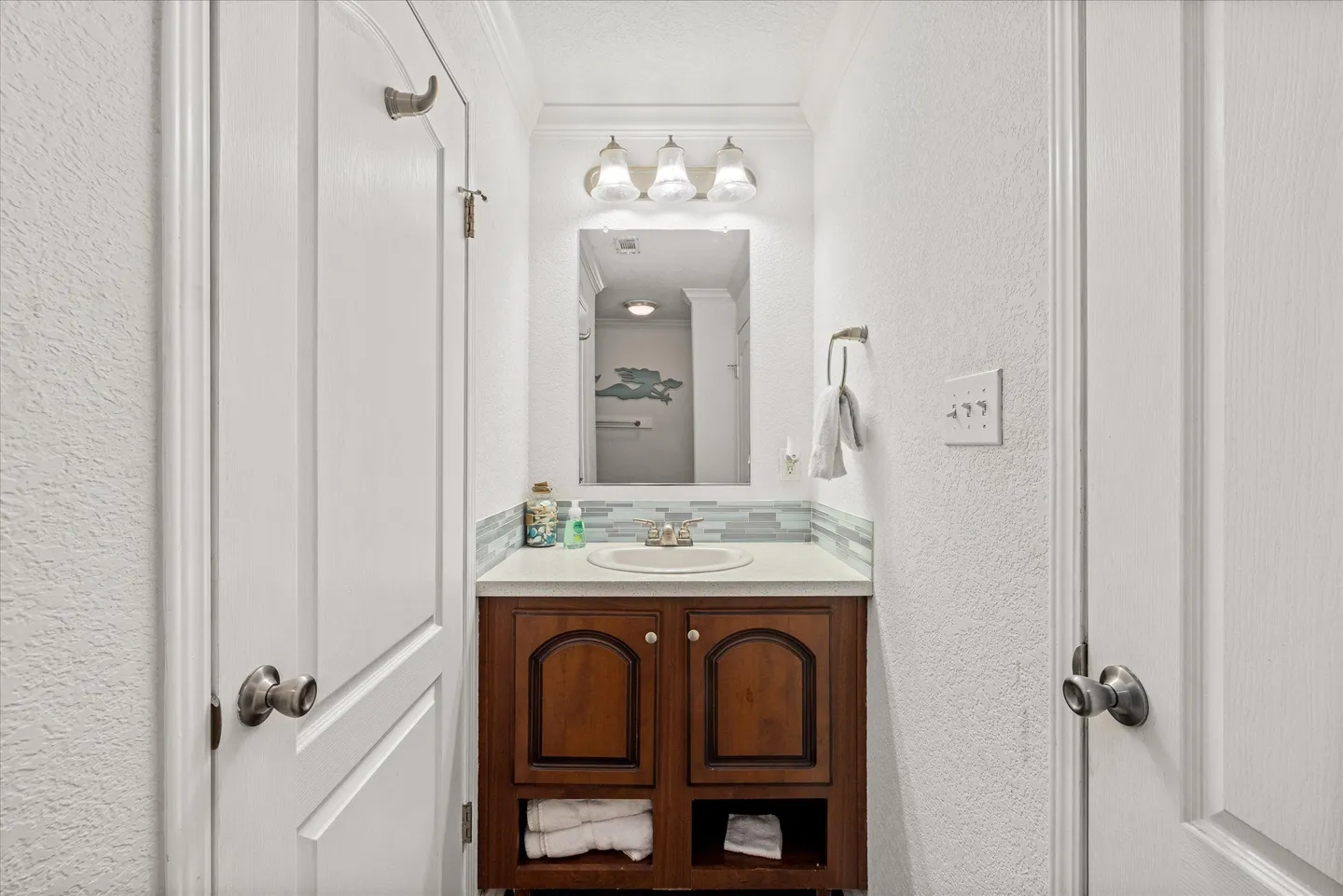 Bathroom vanity framed by open white doors, featuring a dark wood cabinet, white countertop with sink, wall mirror, and vanity lights.