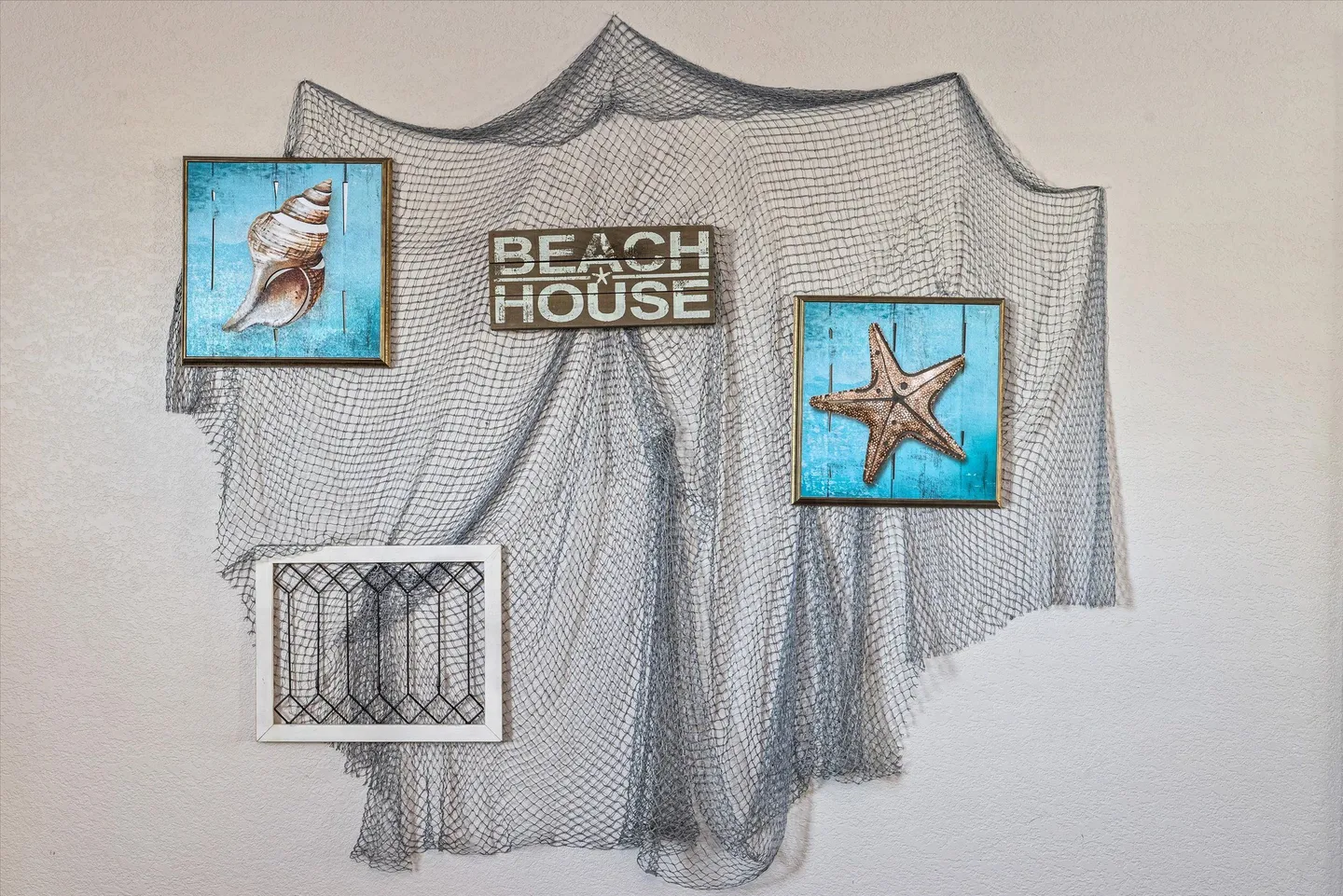 Coastal wall decor featuring a fishing net, a wooden Beach House sign, seashell and starfish artwork, and a framed geometric piece on a white wall.