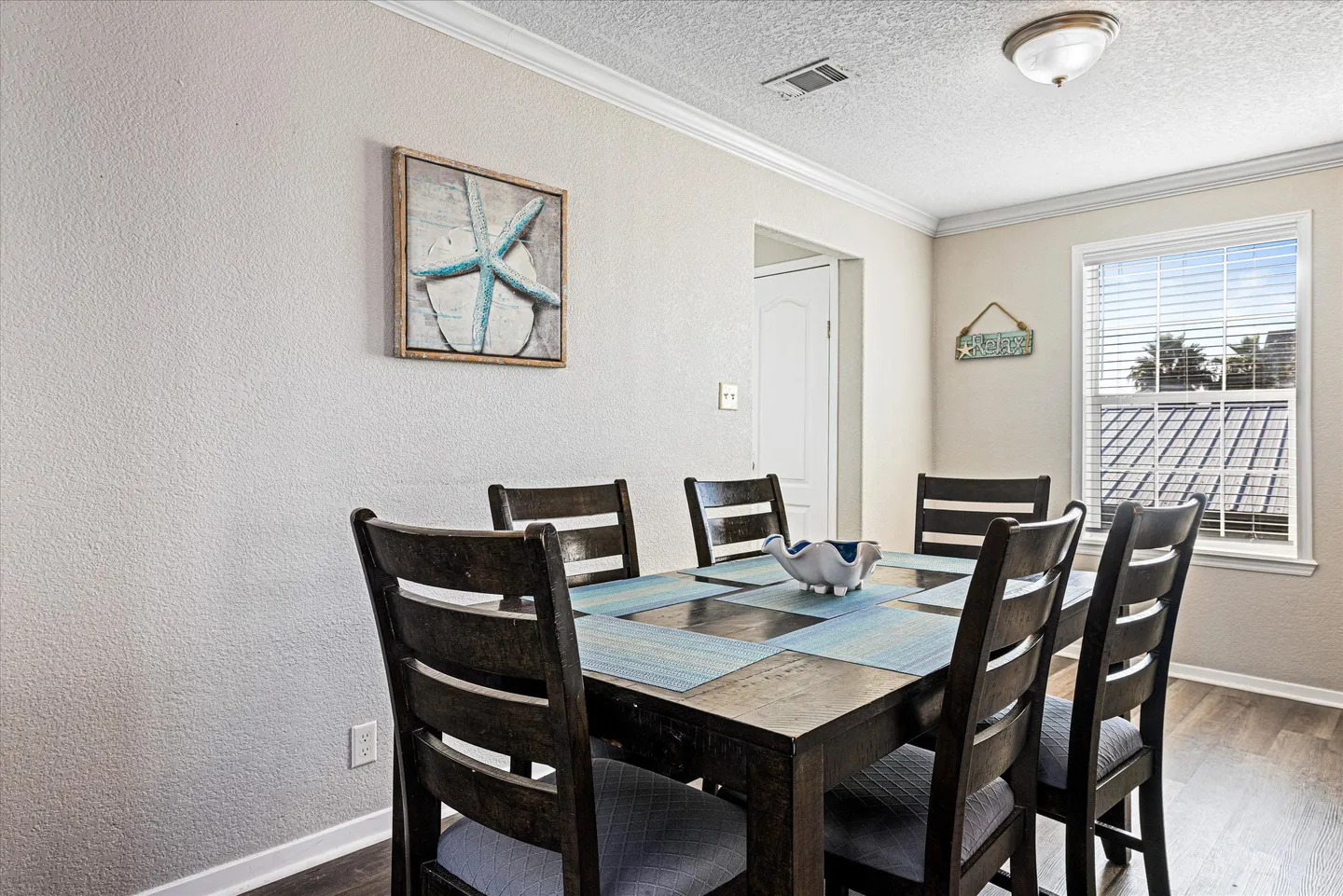 Dining area featuring a dark wooden table with matching chairs, place settings, coastal wall art, and a window with blinds.