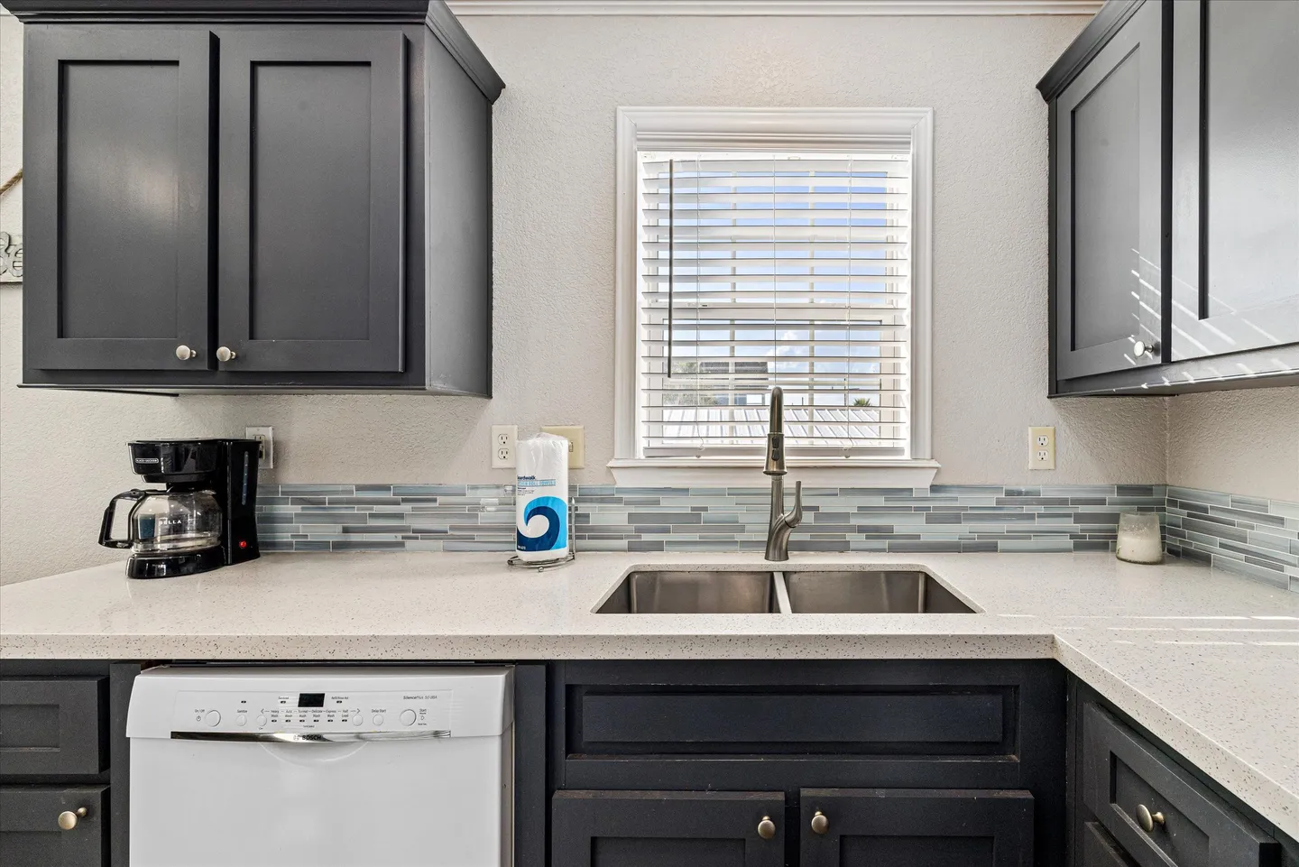 Kitchen counter with dark cabinets, a double sink under a window with blinds, and a white dishwasher.