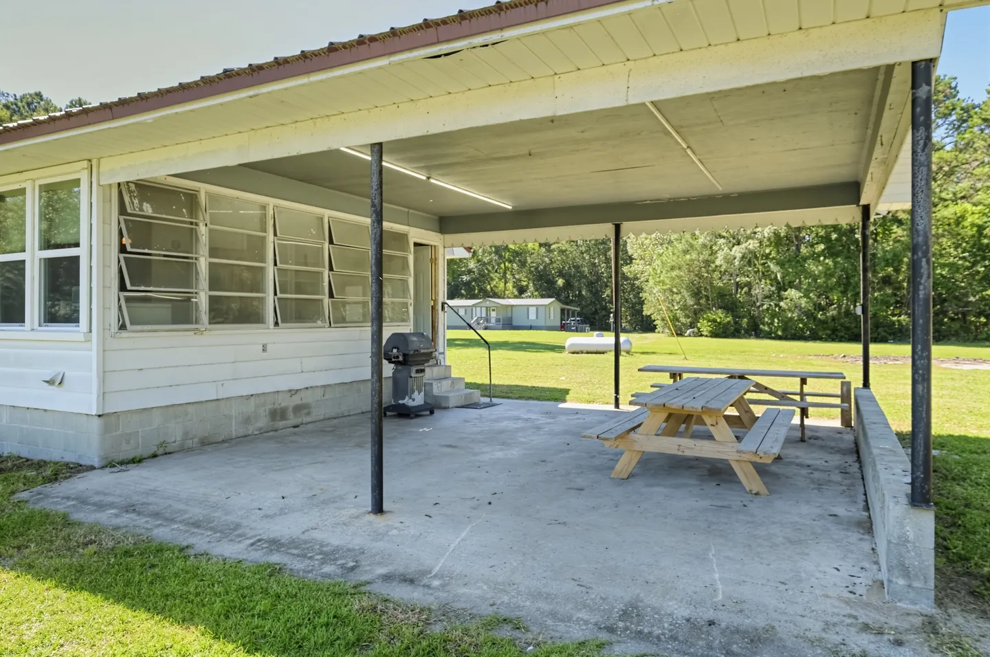 Covered concrete patio with picnic tables, a barbecue grill, and a grassy yard in the background.