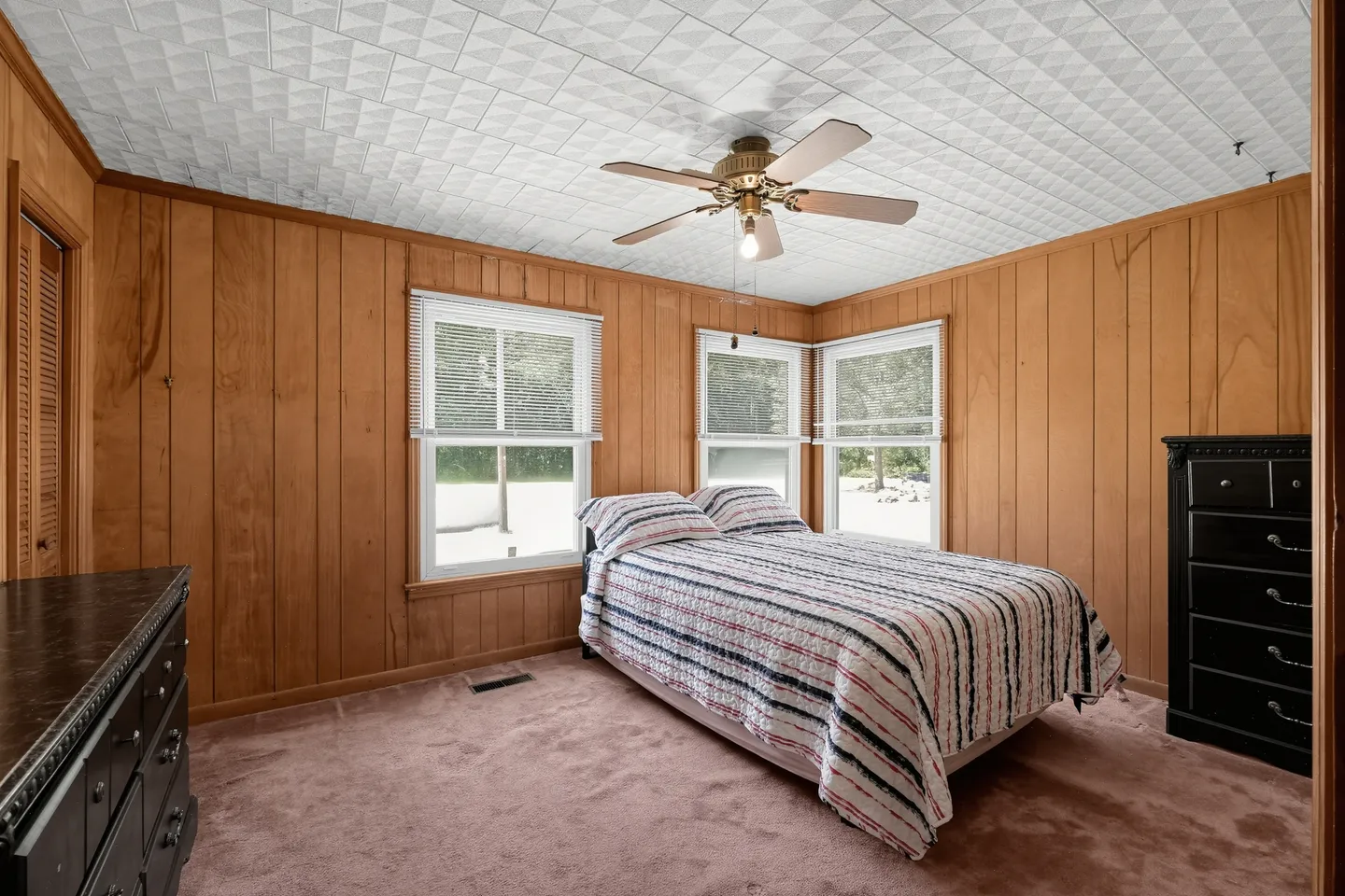 Bedroom with wood-paneled walls, textured white ceiling tiles, carpeted floor, a bed with a striped bedspread, and windows with blinds.