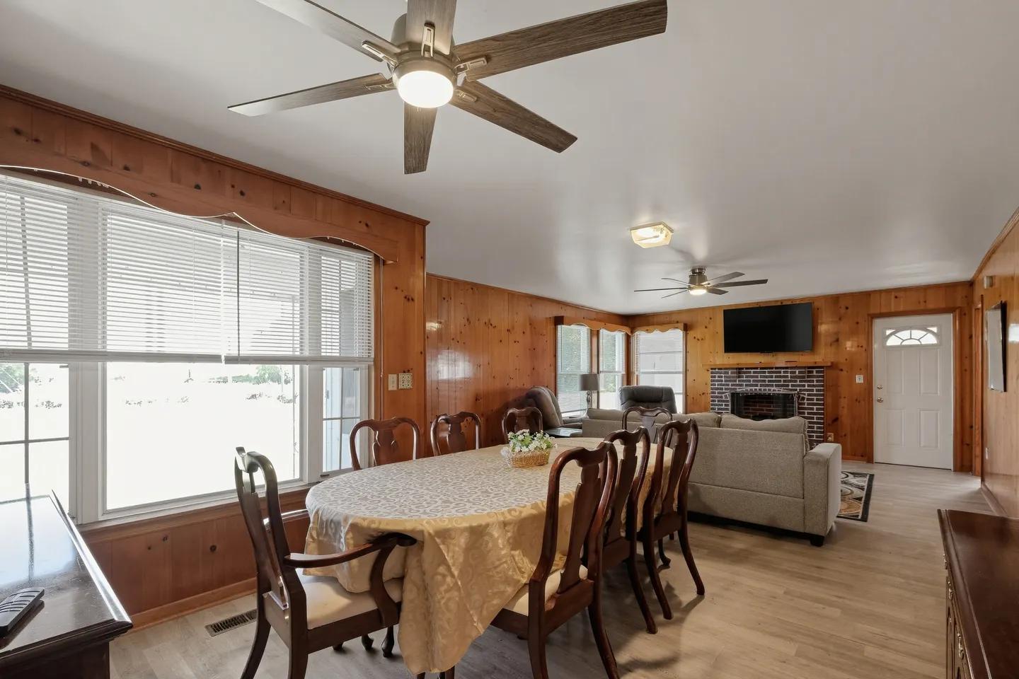 Dining area with an oval table and wooden chairs adjoining an open living room with wood-paneled walls and a brick fireplace.