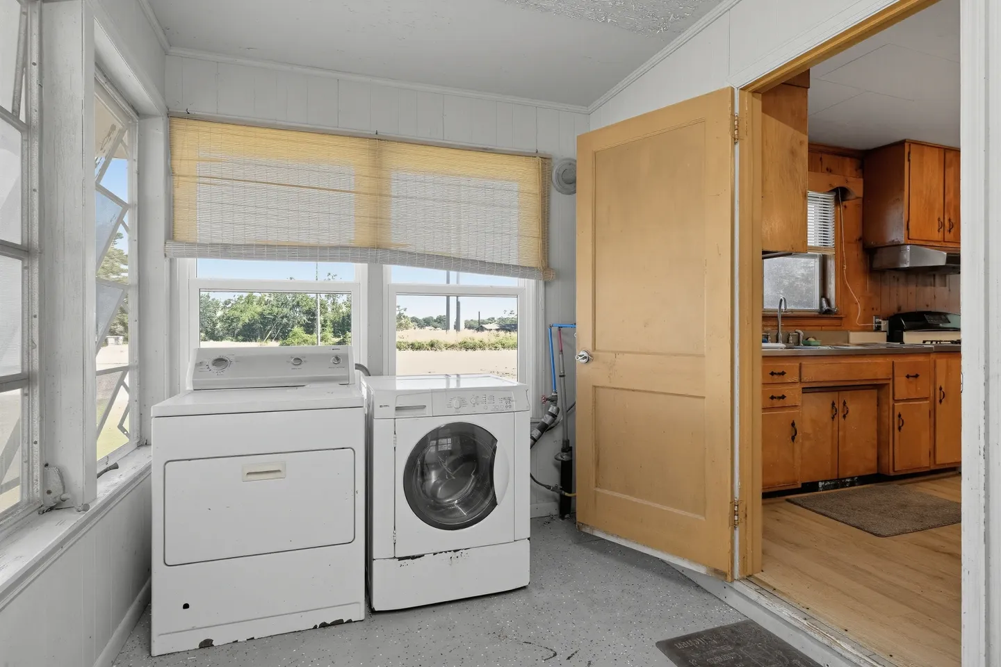 Sunroom laundry area with a white washing machine and dryer next to windows, leading into a kitchen with wood cabinets.