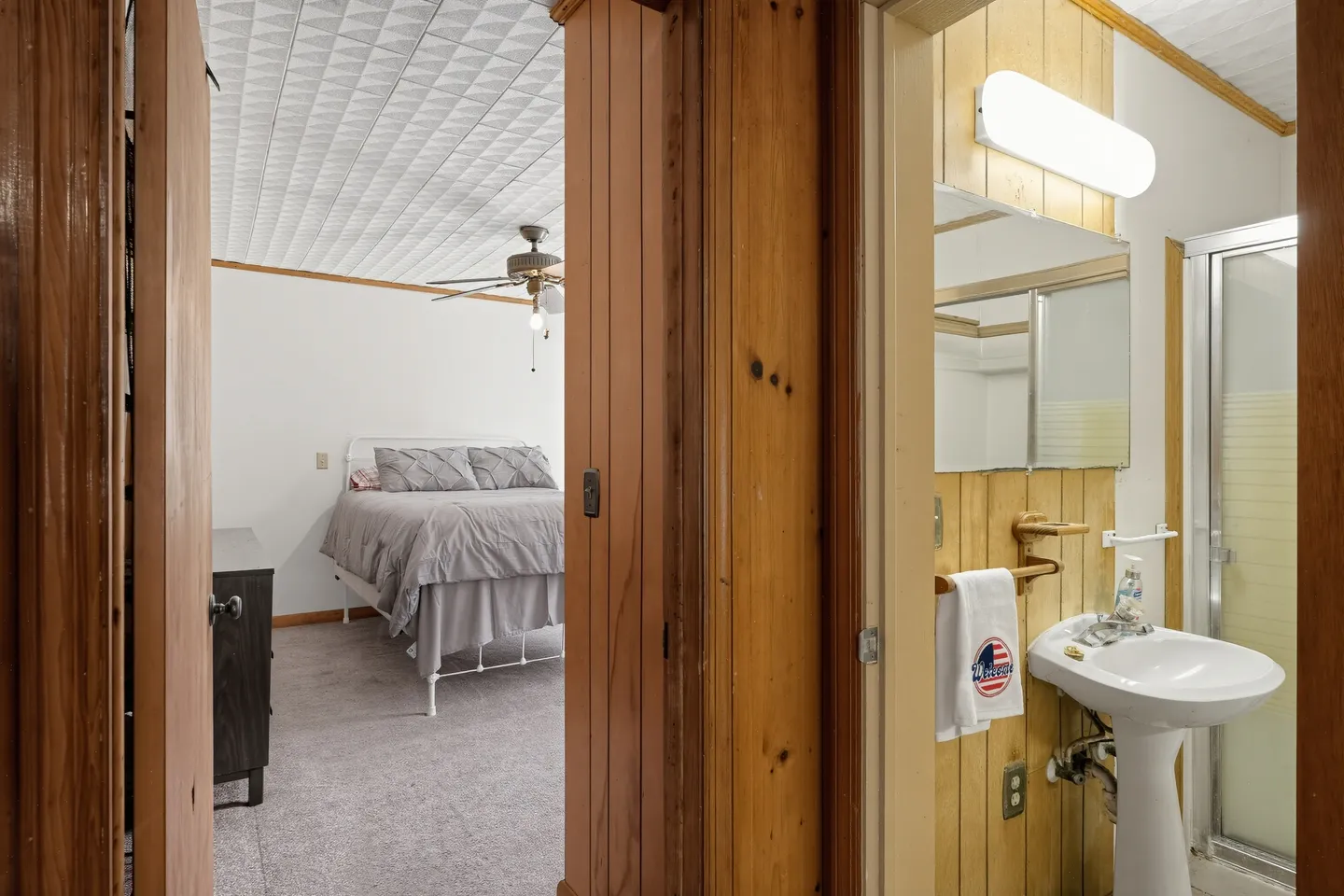 A split view from a wood-paneled hallway showing an adjoining bedroom with a made bed and a bathroom with a pedestal sink.