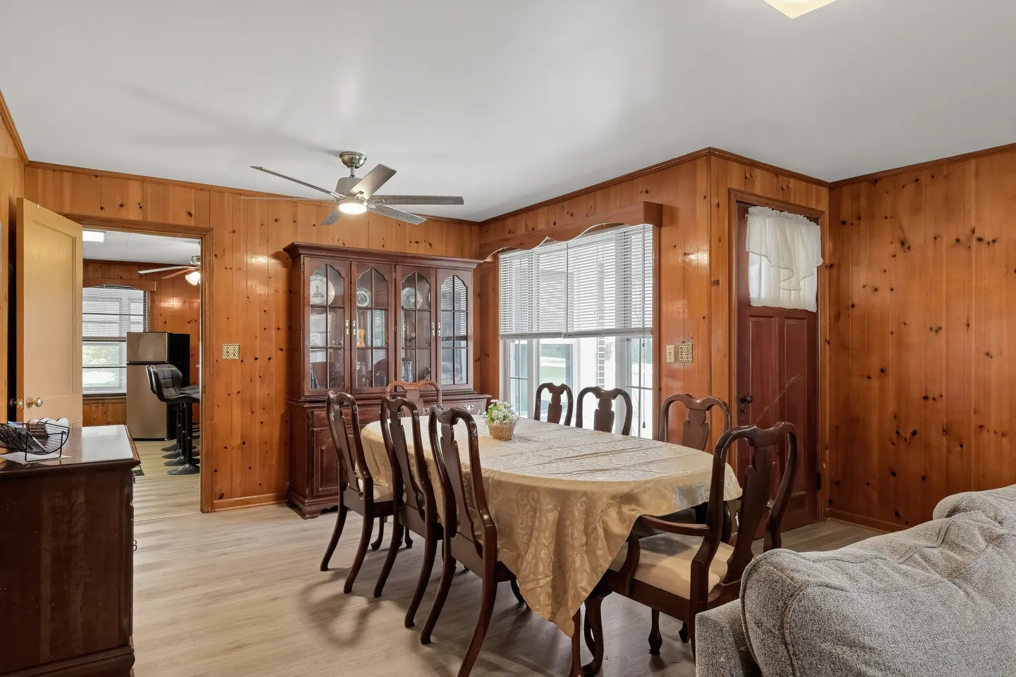 Dining area with wood-paneled walls, a large dining table with chairs, a wooden china cabinet, and a view into the kitchen.