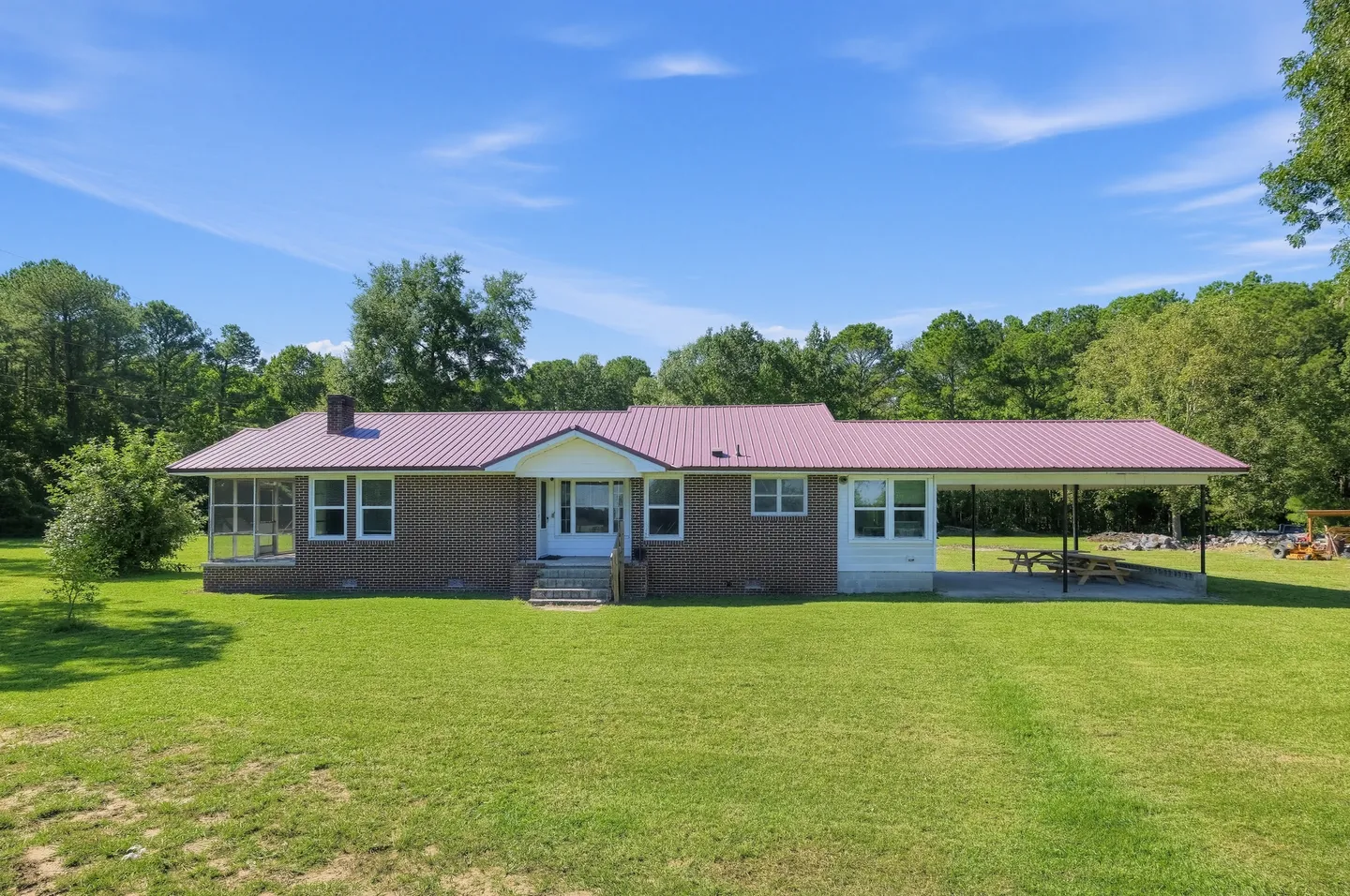 Exterior front view of a single-story brick ranch home with a reddish-pink metal roof, carport, and wide grassy yard under a clear blue sky.