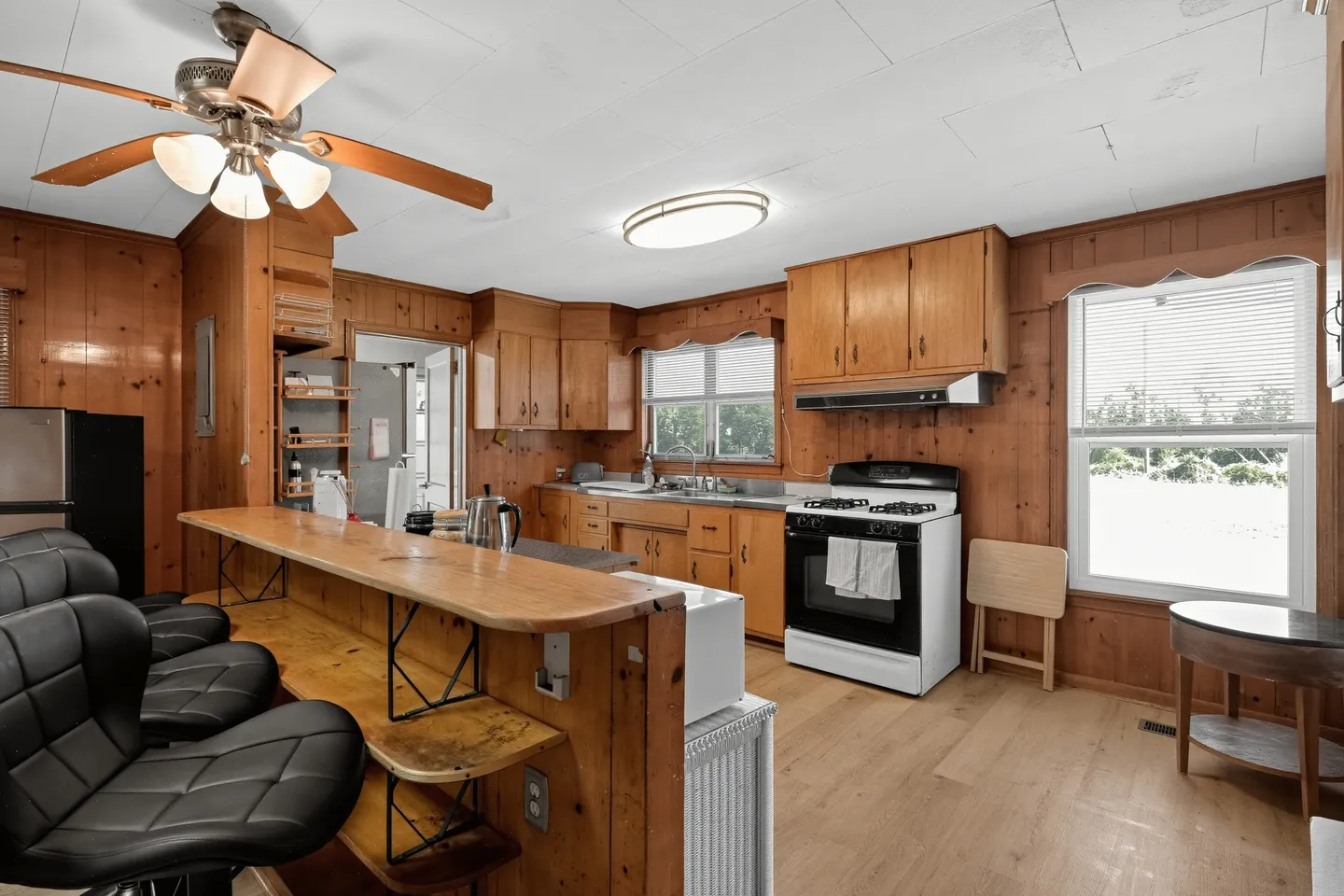 Kitchen with wood-paneled walls and cabinets, a breakfast bar with black stools, white appliances, and a ceiling fan.