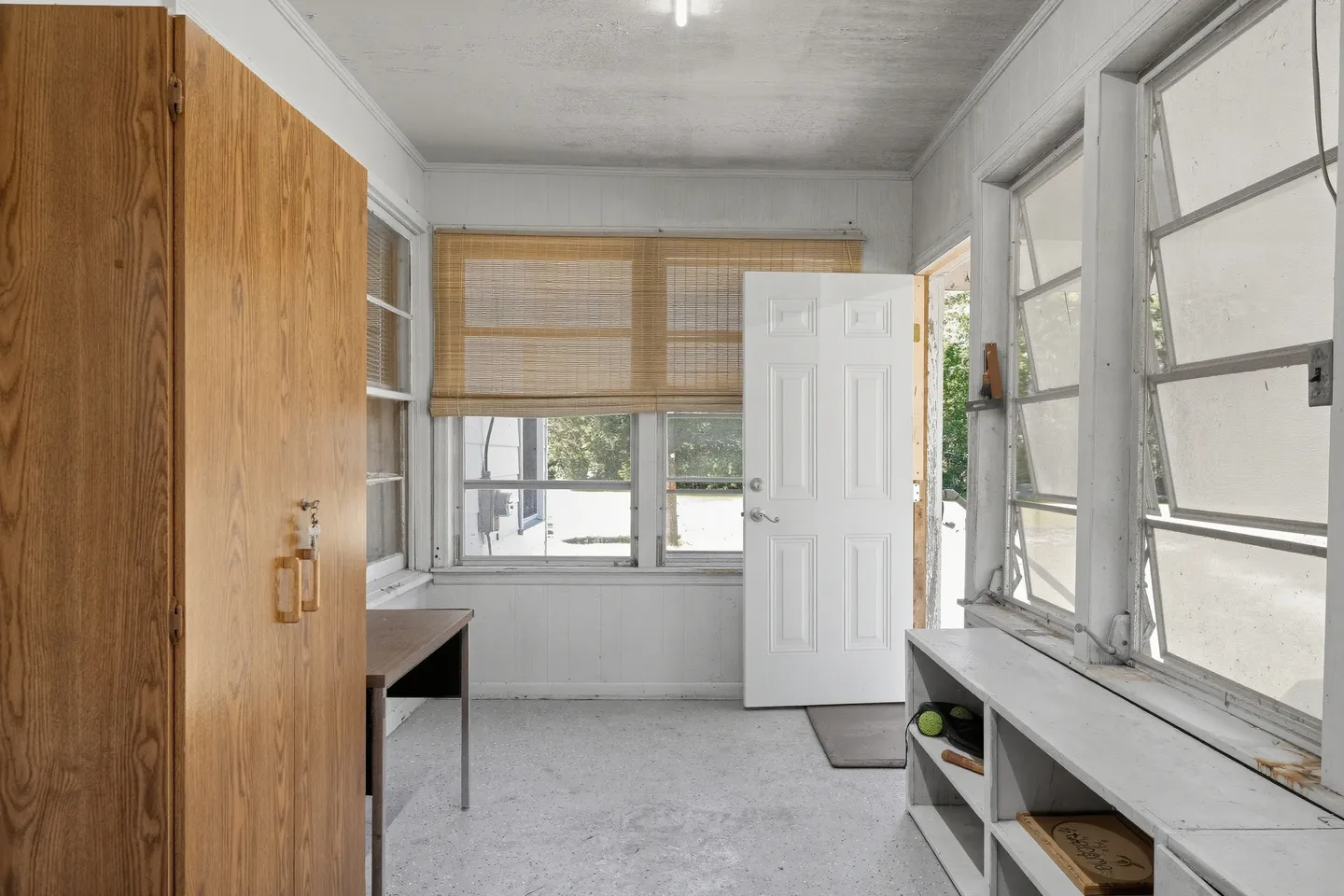 Sunroom or utility porch with white walls, multiple windows, a tall wooden cabinet, low white shelving, and a white exterior door.