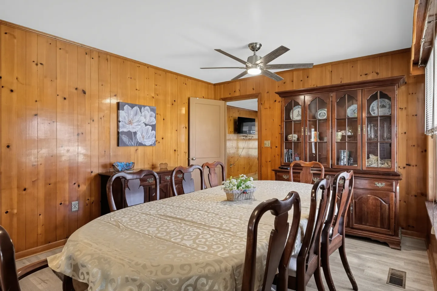 Dining room with wood paneled walls, a large dining table covered with a patterned tablecloth, matching wooden chairs, and a glass-front china cabinet.