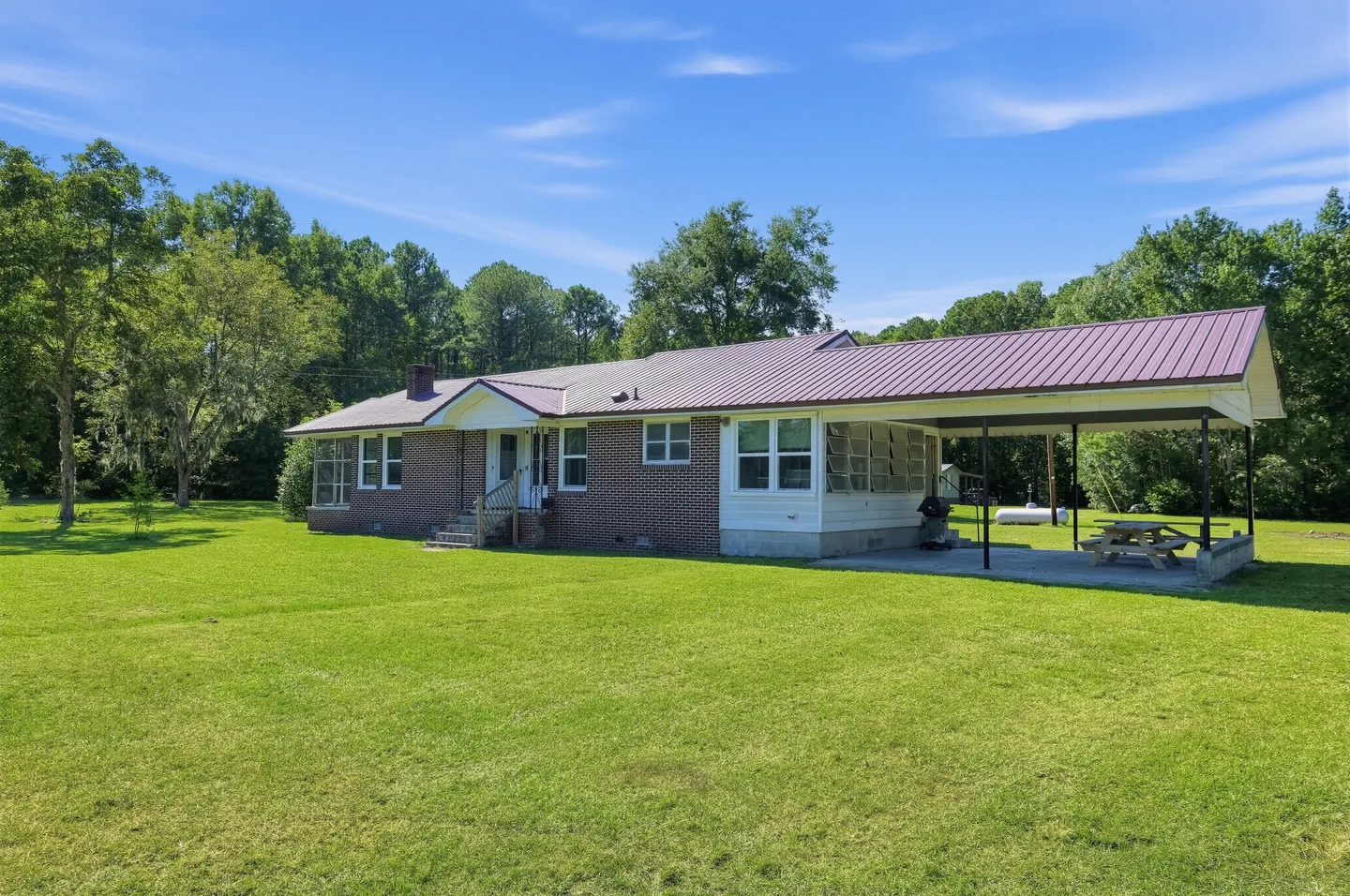 Exterior of a single-story brick home with a maroon metal roof, covered carport, and large grassy lawn under a blue sky.