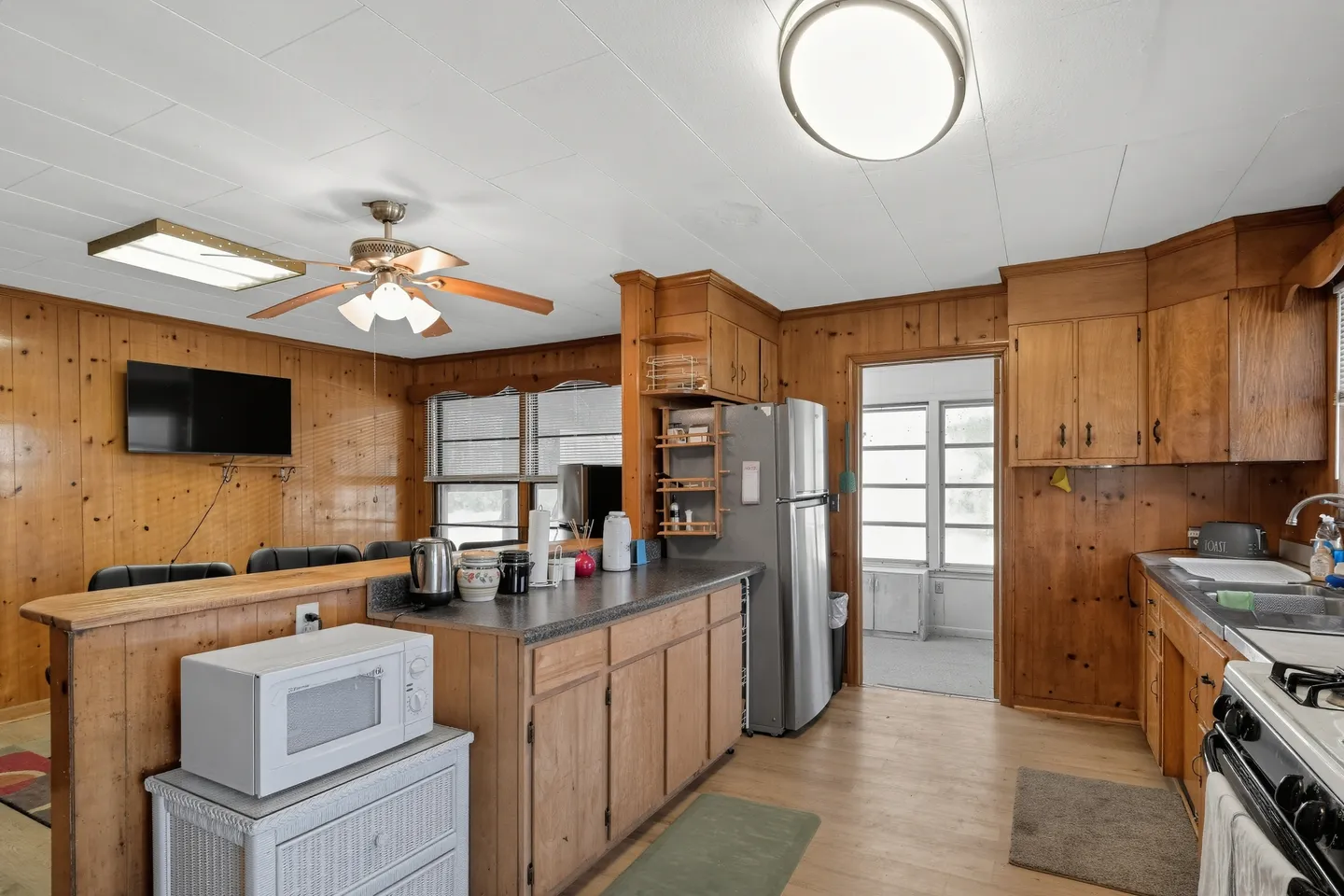 Kitchen with wood-paneled walls and cabinets, stainless-steel refrigerator, white ceiling tiles, and a view into an adjacent sunroom.
