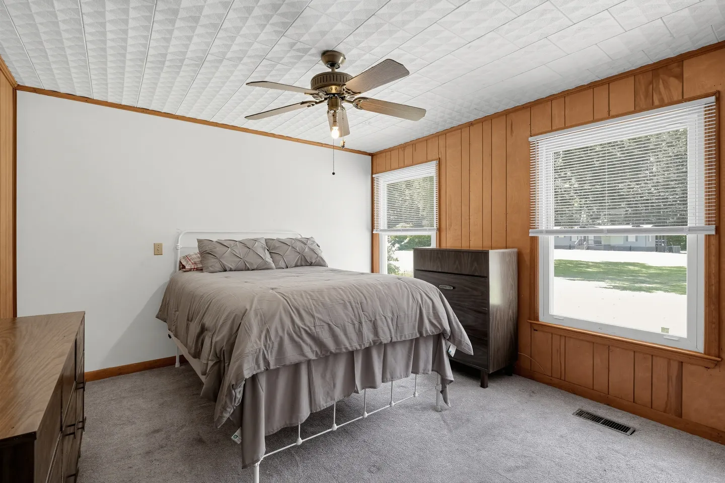 Bedroom with carpet, wood-paneled wall, textured ceiling, a bed with gray bedding, a ceiling fan, and two windows.