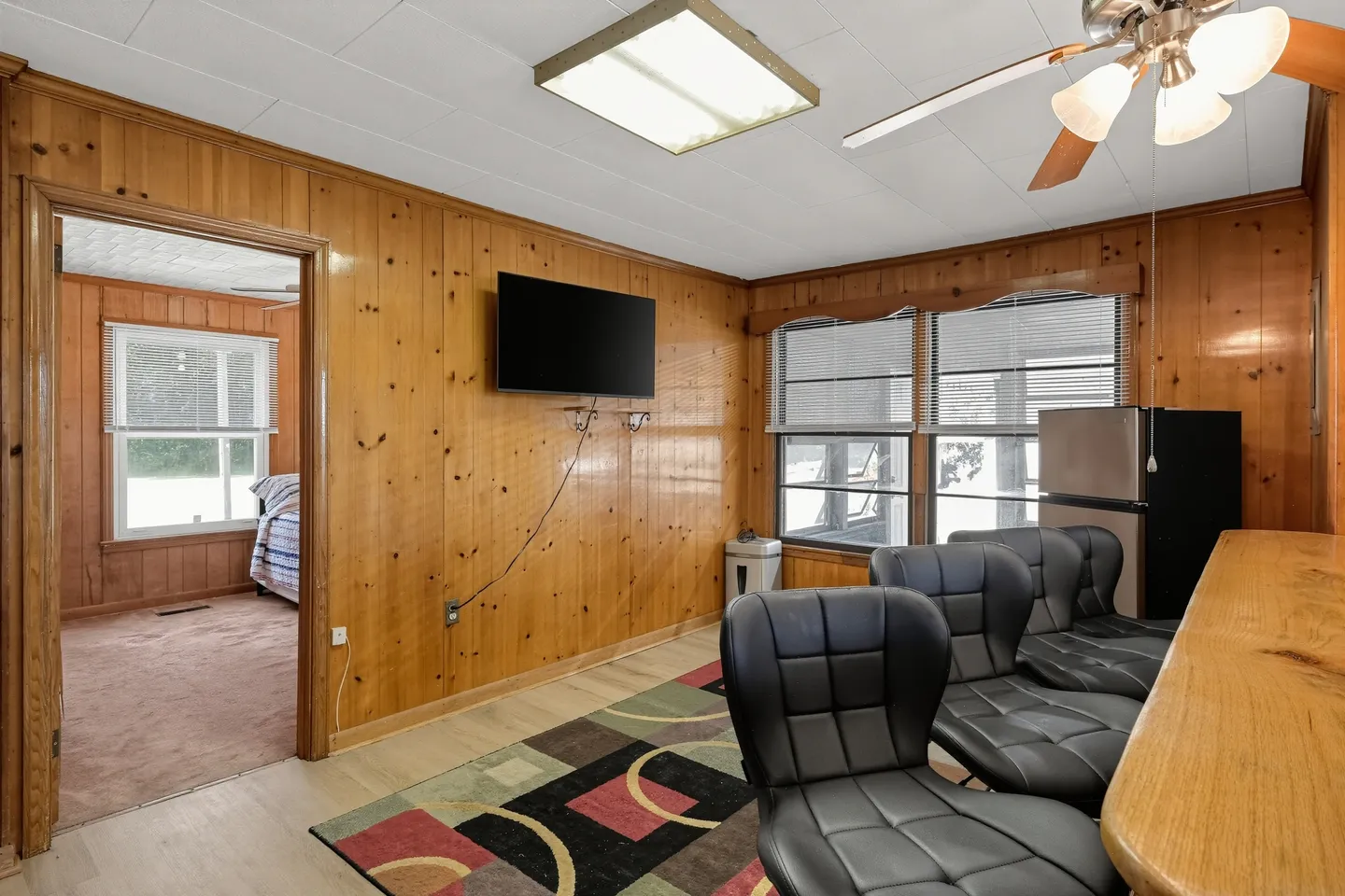 Living or recreation room with wood paneled walls, a wall-mounted TV, several black swivel chairs along a counter, and an open doorway showing a bedroom.