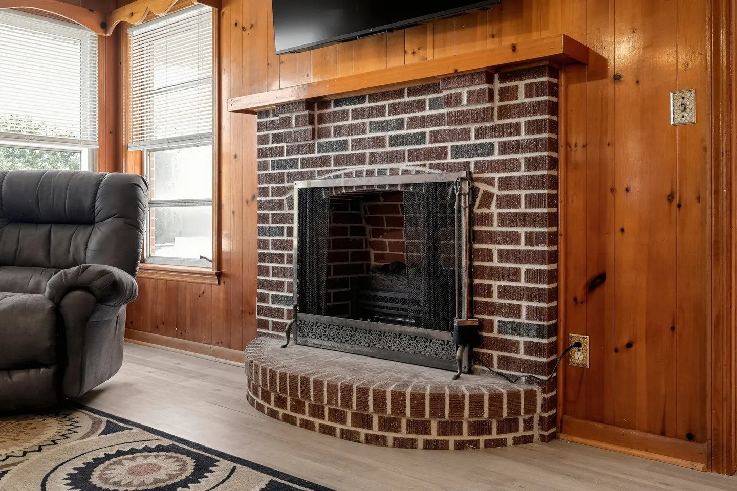 Brick fireplace with a curved hearth, wood mantel, and screen in a wood-paneled room.