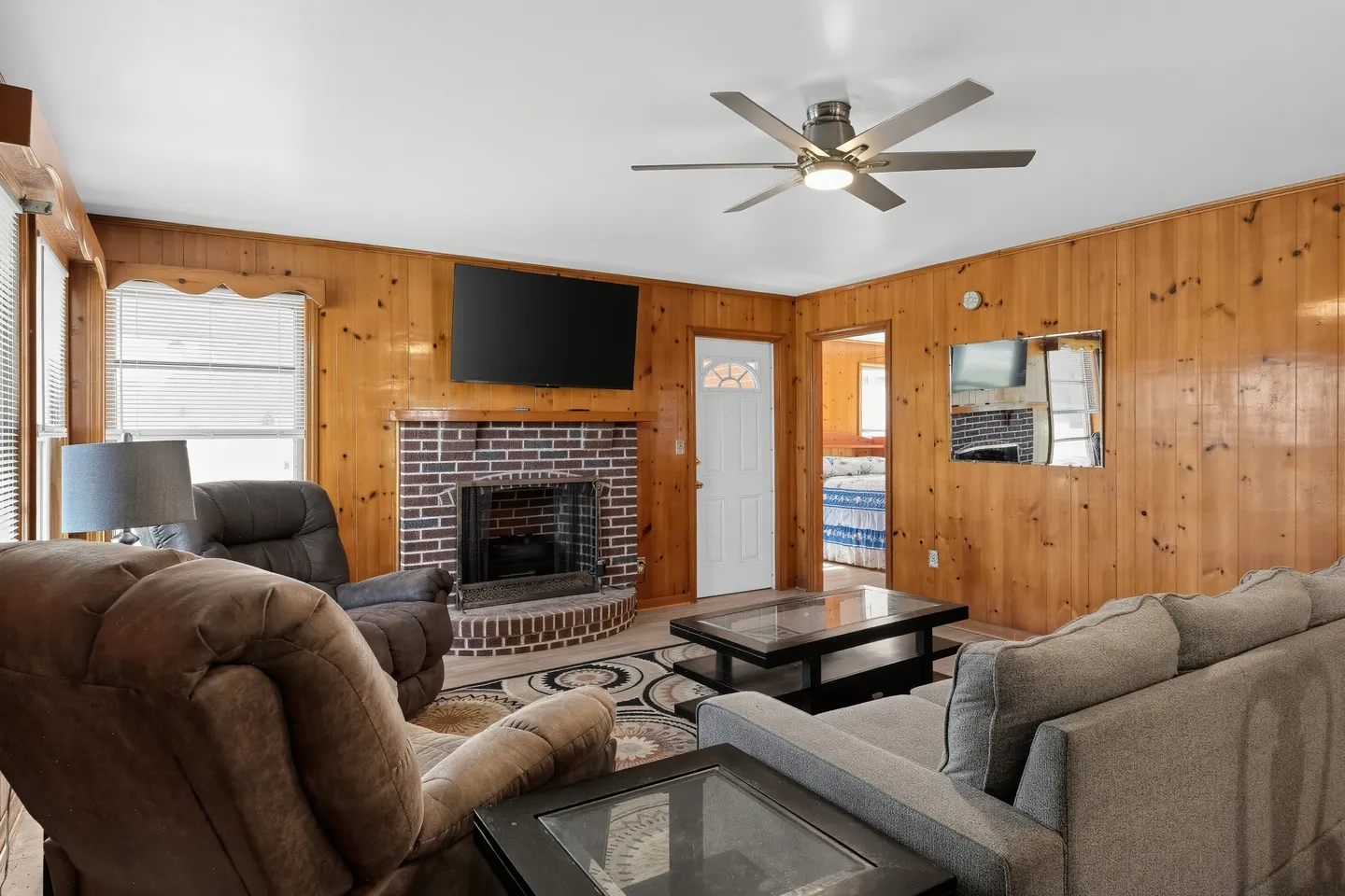 Living room featuring wood-paneled walls, a brick fireplace with a mounted television, a ceiling fan, and seating including a sofa and armchairs.