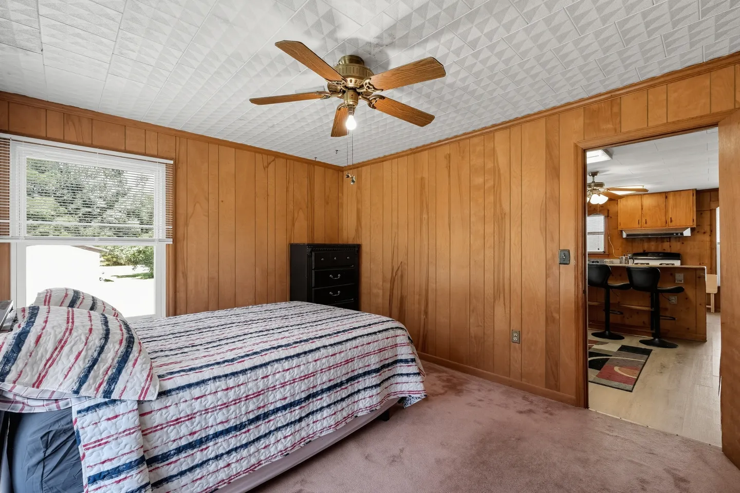 Bedroom with wood-paneled walls, carpeted floor, ceiling fan, and a view into the adjacent kitchen.