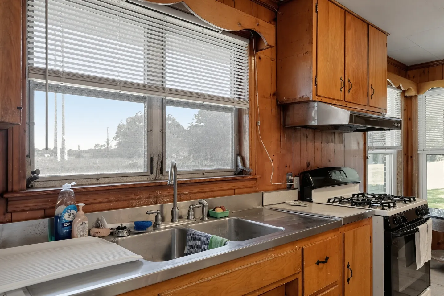 Kitchen counter with a double stainless steel sink below a window with white blinds, flanked by wood-paneled walls and cabinets.