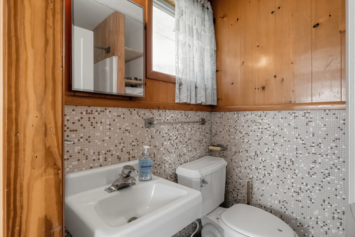 Bathroom with wood-paneled upper walls, mosaic tile wainscoting, a wall-mounted sink, toilet, and mirrored medicine cabinet.