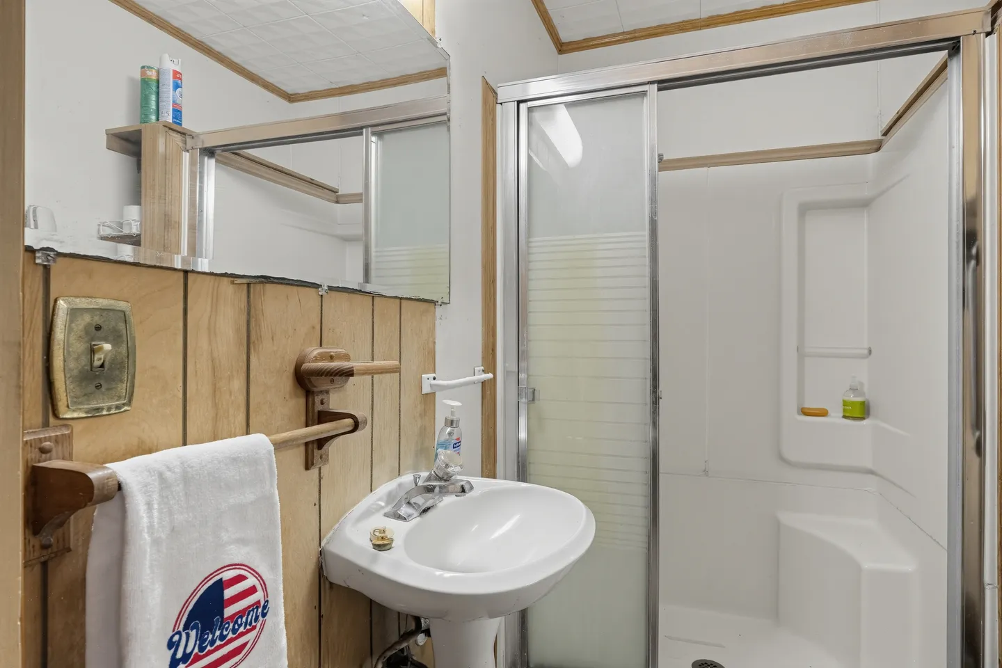 Bathroom with a pedestal sink, wood-paneled wall, mirror, and a walk-in shower stall with sliding glass doors.