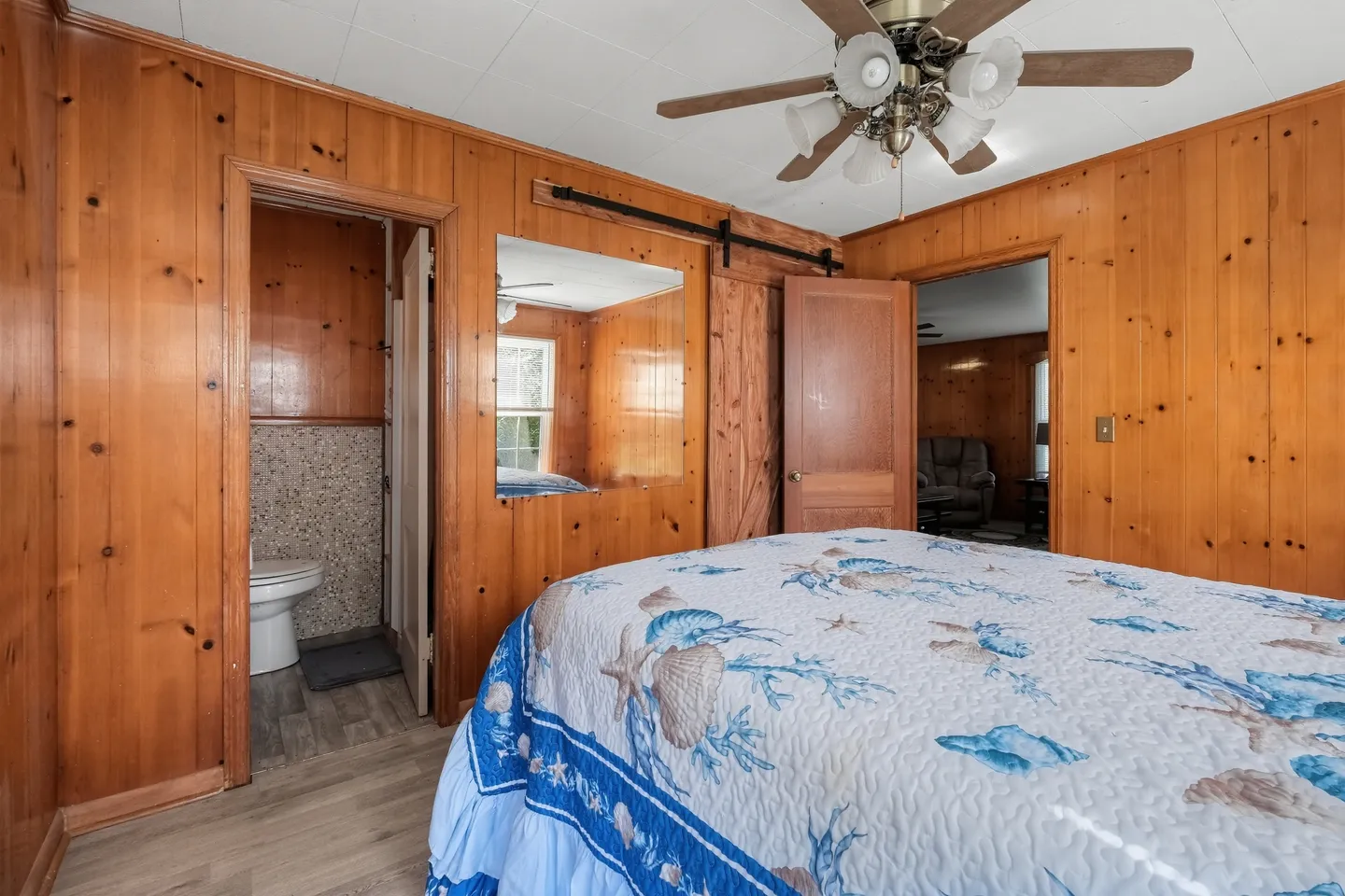 Bedroom with wood-paneled walls, a ceiling fan, a bed with a blue seashell patterned quilt, and an open doorway to an en-suite bathroom.