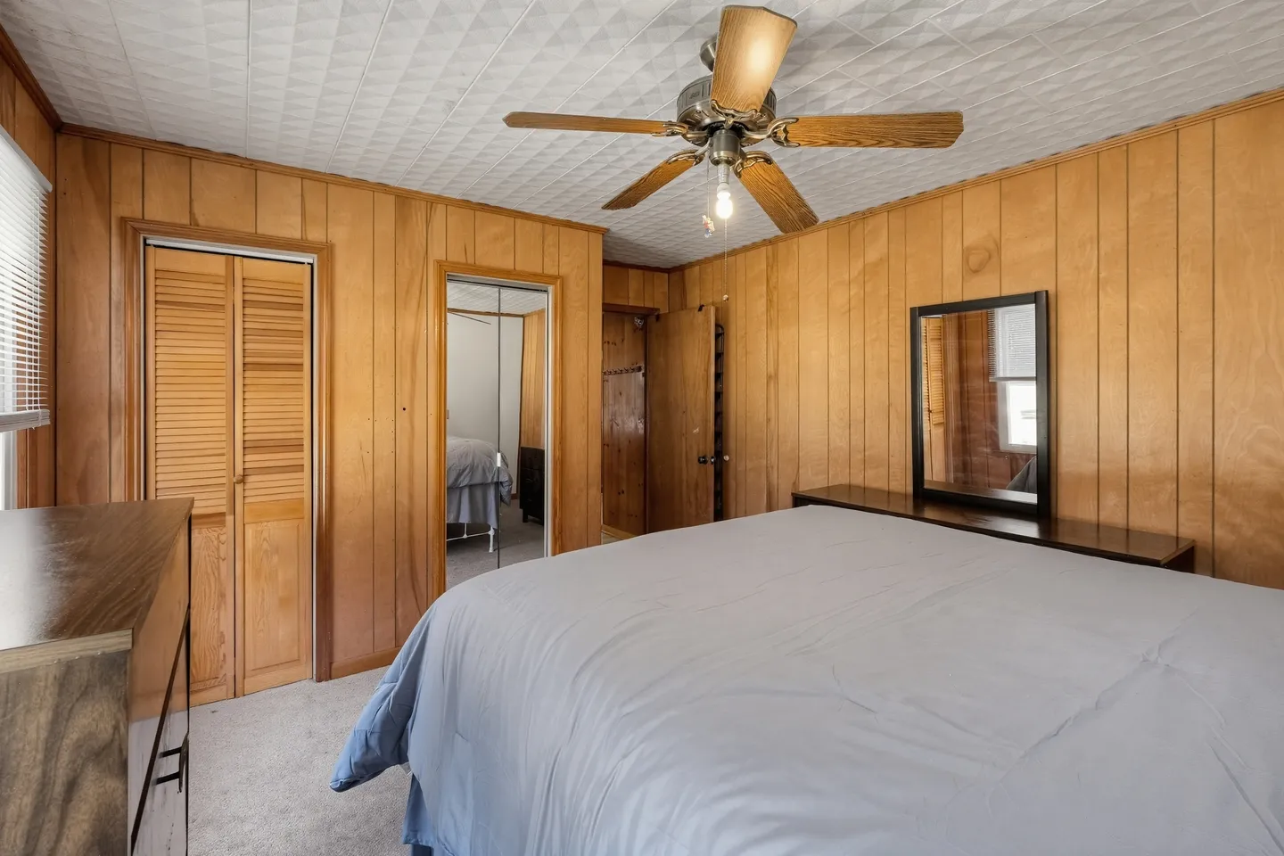 Bedroom featuring wood panel walls, textured ceiling with a fan, a bed made with blue bedding, and a mirrored closet door.