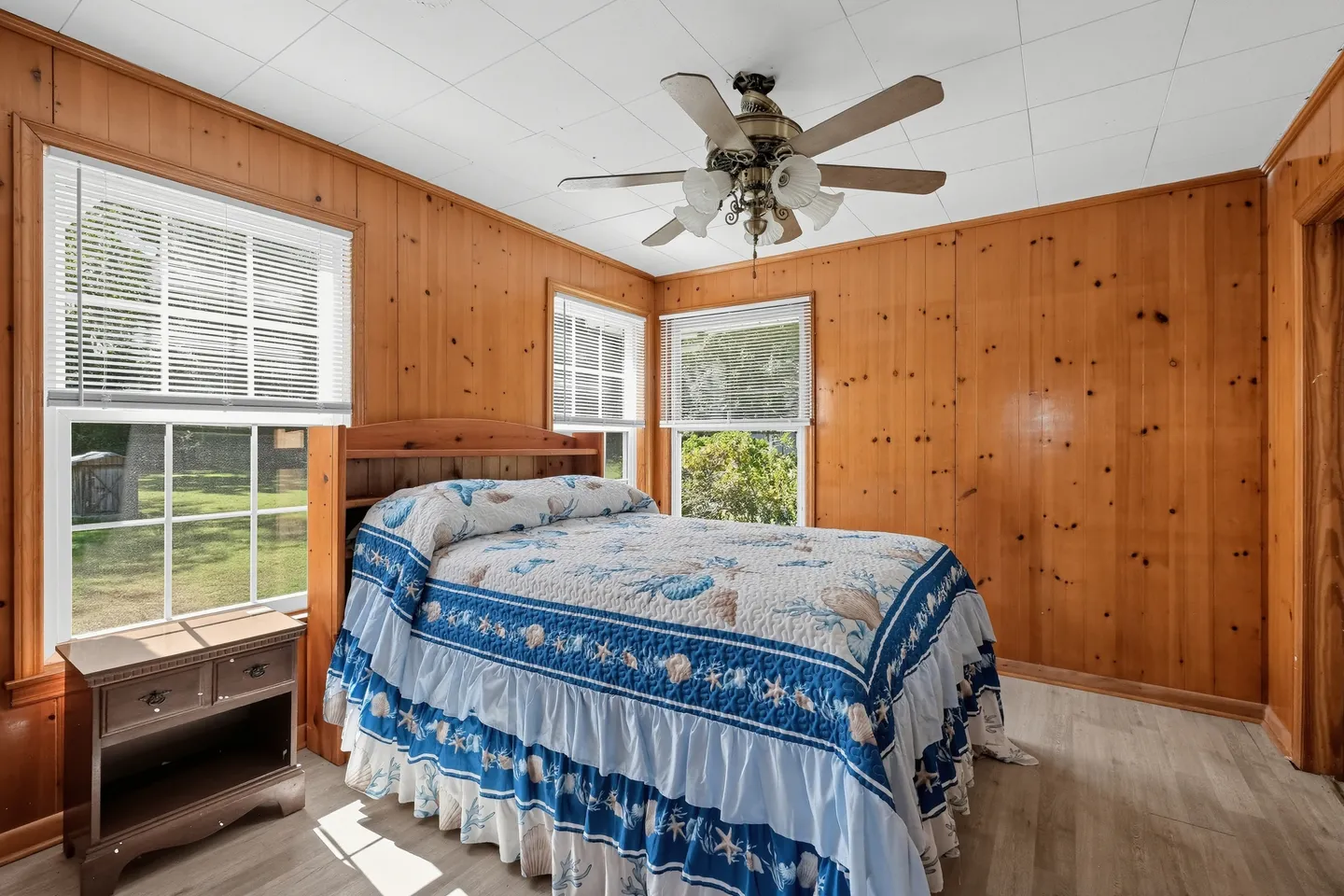 Bedroom with knotty pine wood-paneled walls, a bed with a blue and white coastal-themed quilt, and multiple windows with blinds.