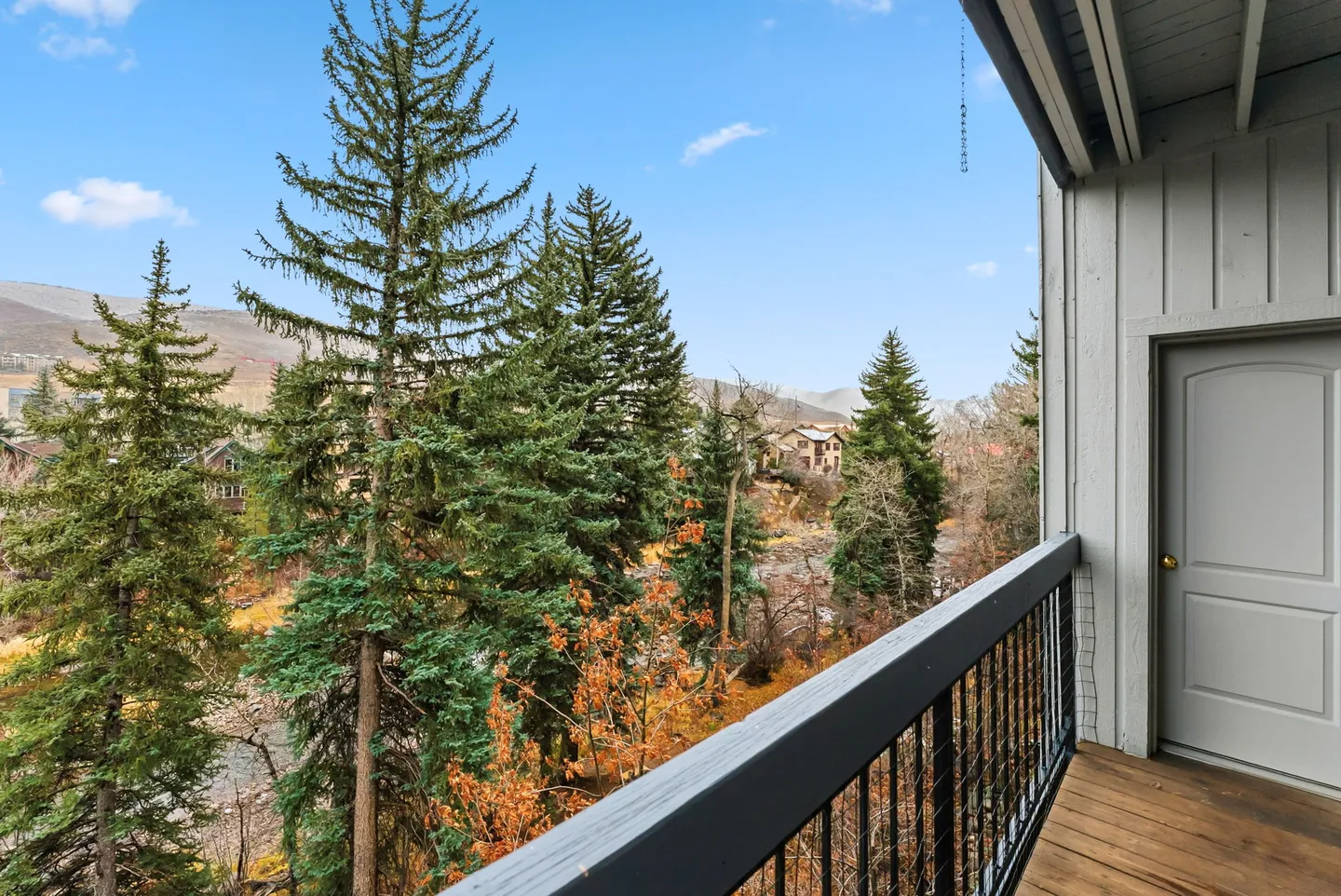 Elevated view from a wooden deck with dark railings overlooking tall evergreen trees, distant houses, and hills under a blue sky.