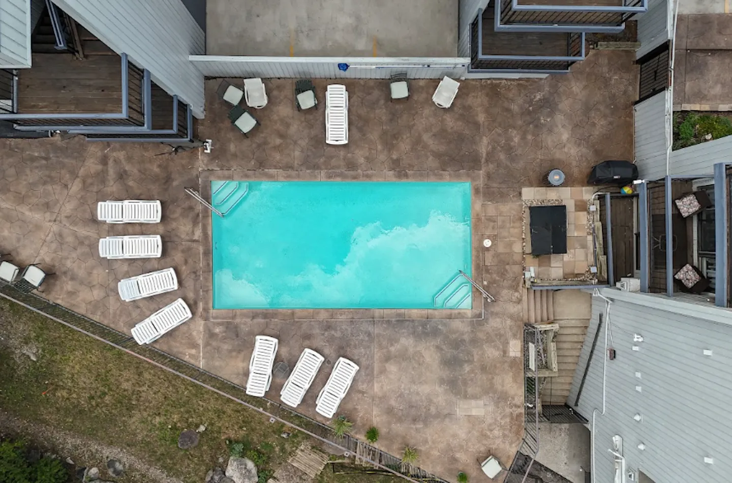 Overhead view of a rectangular outdoor swimming pool surrounded by a patio with lounge chairs and adjacent apartment or condo buildings.