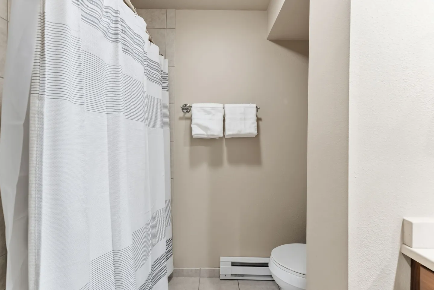 Bathroom showing a striped shower curtain, folded white towels on a wall rack, a baseboard heater, and a partial view of a toilet.