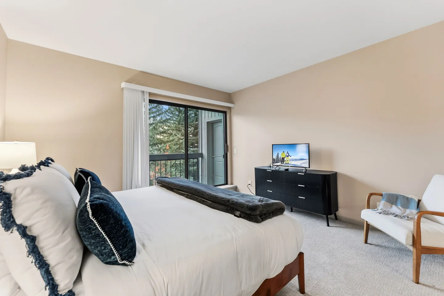 Bedroom with a made bed, sliding glass door leading to a balcony, dark dresser with a television, and an accent armchair.
