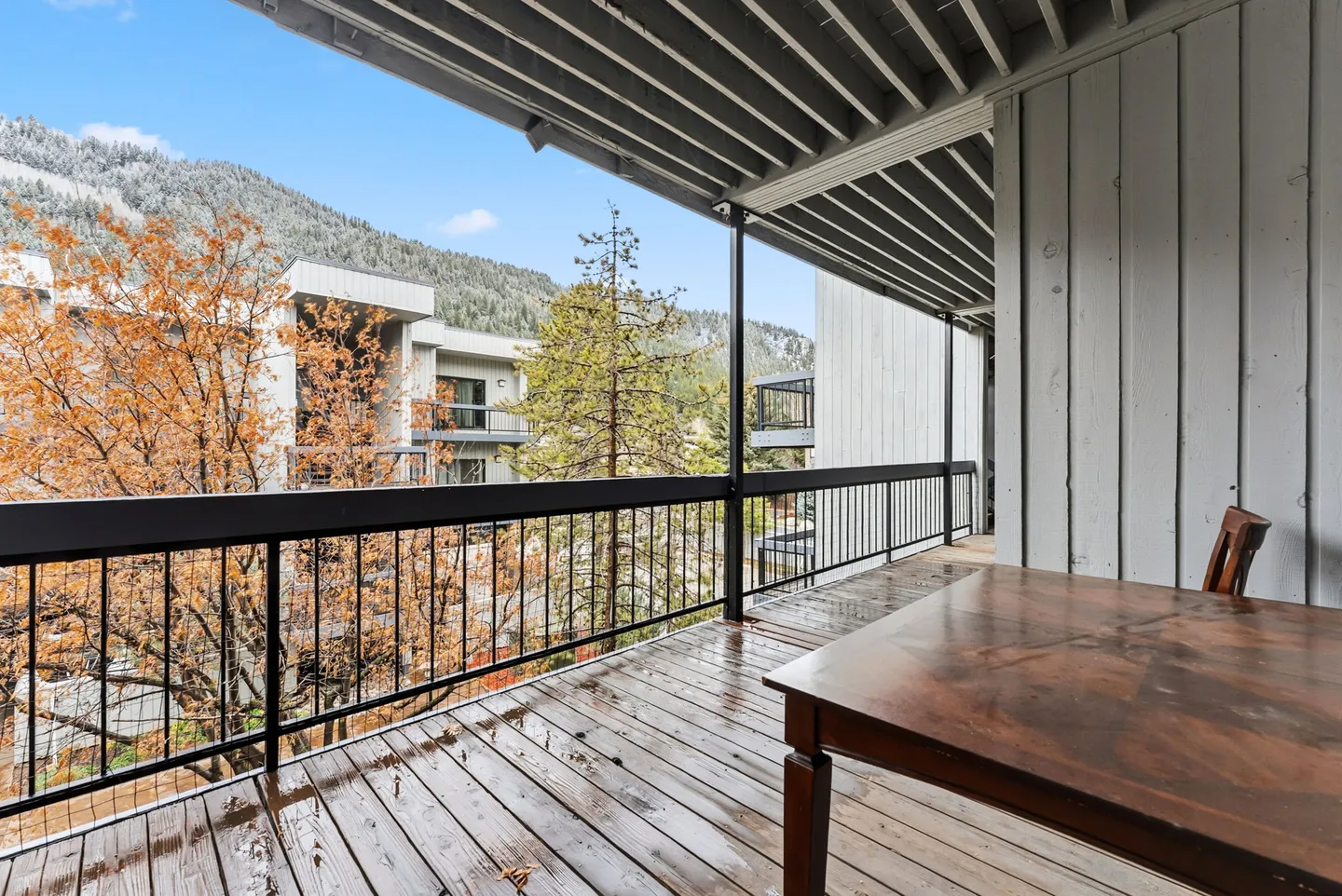 Covered outdoor balcony with a wooden floor, railing, table, and chair looking out toward trees and mountains.