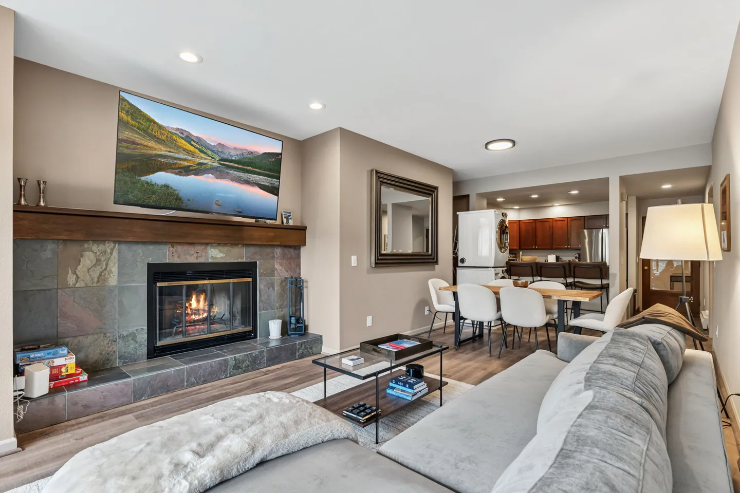 Open living area with a fireplace, TV mounted above, gray sectional sofa, dining table, and kitchen in the background.