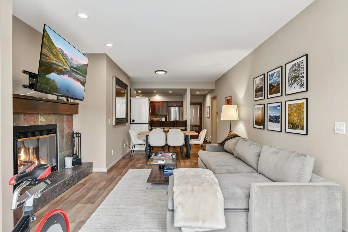 Open-concept living room with a fireplace, wall-mounted TV, sectional sofa, and adjacent dining area leading to the kitchen.