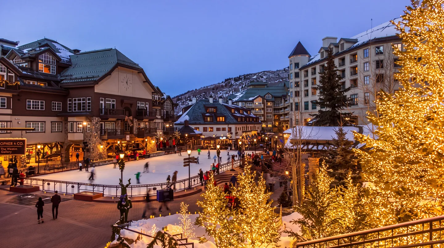 Elevated view of an outdoor ice skating rink surrounded by illuminated alpine-style resort buildings and trees with lights at dusk.
