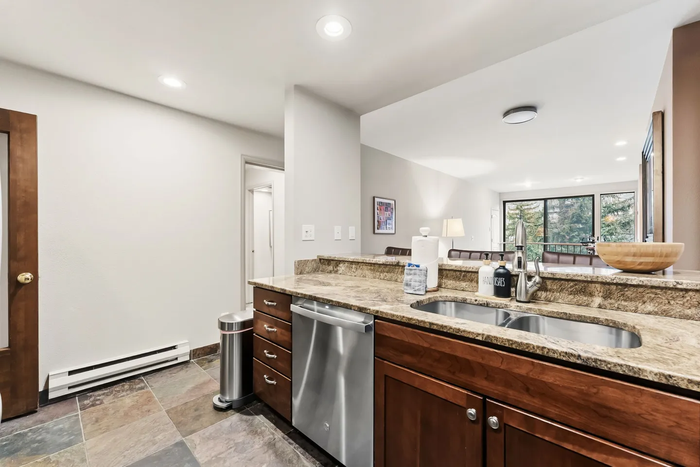 Kitchen counter with a double sink, dishwasher, and wood cabinets overlooking an open living area.
