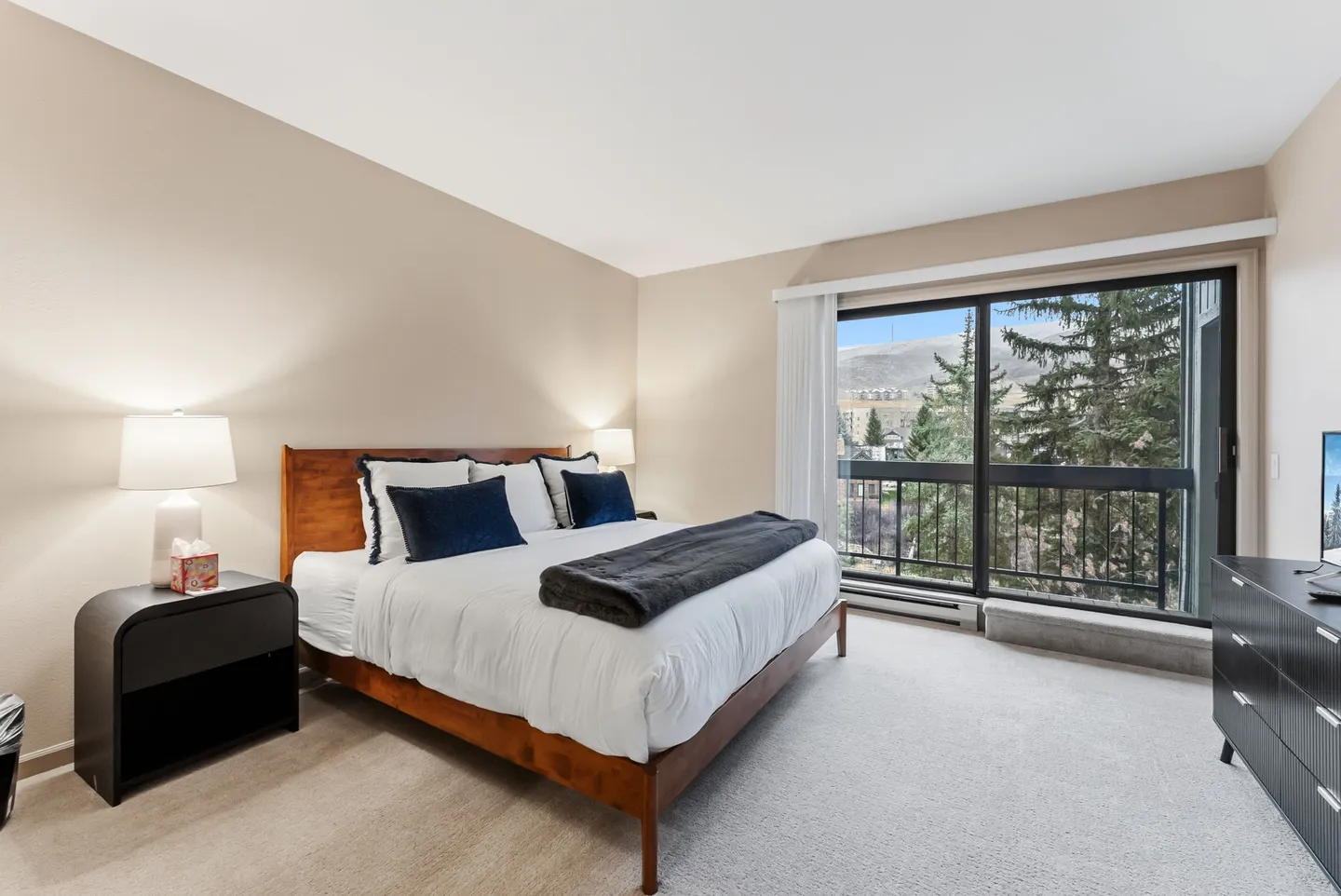 Bedroom with a wooden frame bed, matching nightstand, and a large sliding glass door showing trees and a hillside outside.