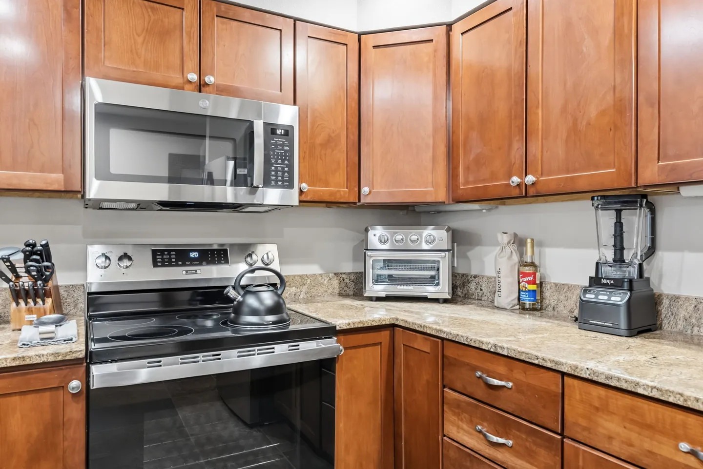 Kitchen corner featuring wooden cabinets, speckled granite countertops, an electric stove, over-the-range microwave, toaster oven, and blender.